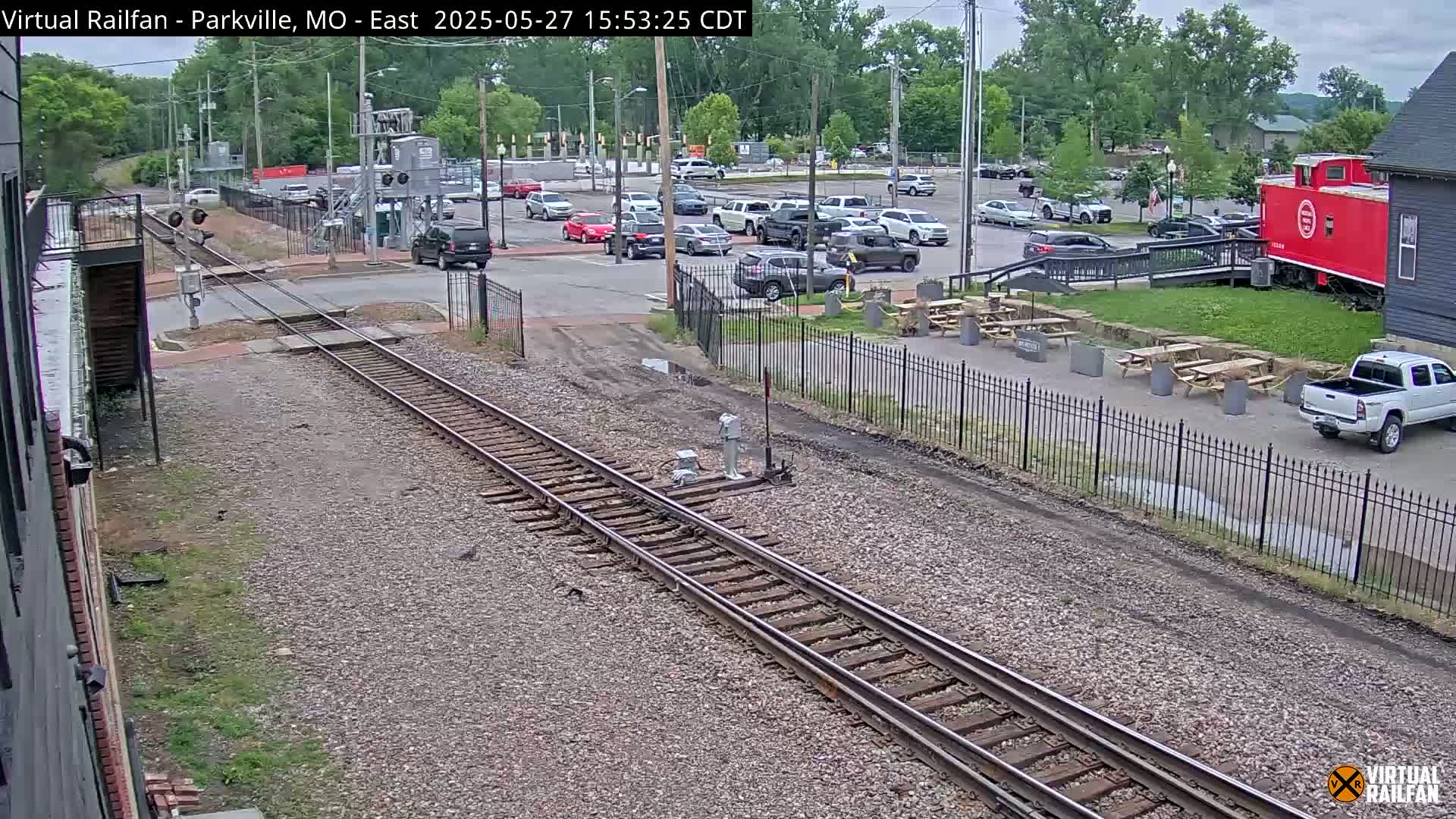A view of railroad tracks and a parking lot with several cars, a red caboose, and picnic tables under an overcast sky.
