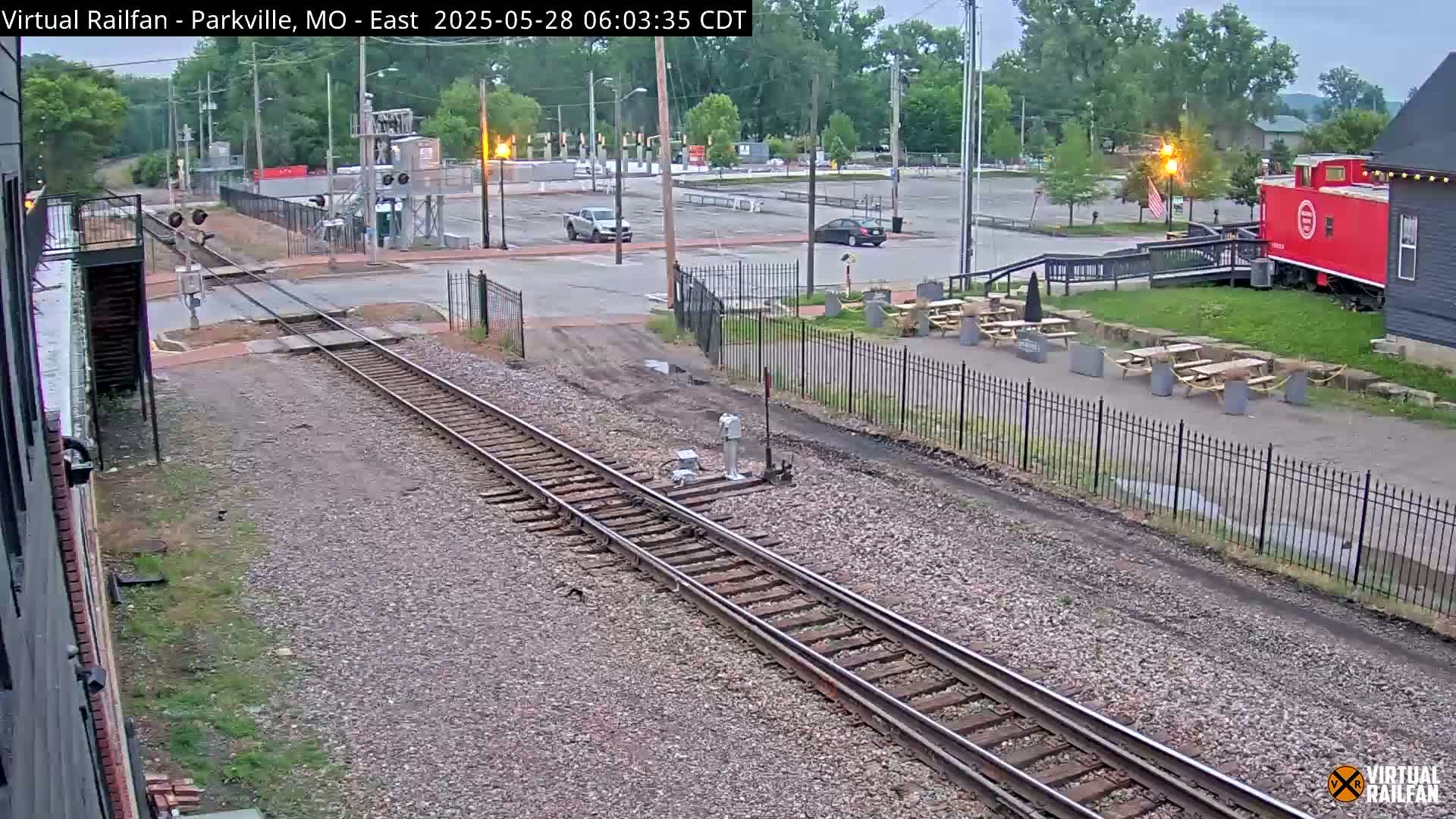 A high-angle view shows railroad tracks intersecting a road next to an outdoor seating area with picnic tables, a parking lot, and a red caboose under an overcast sky.