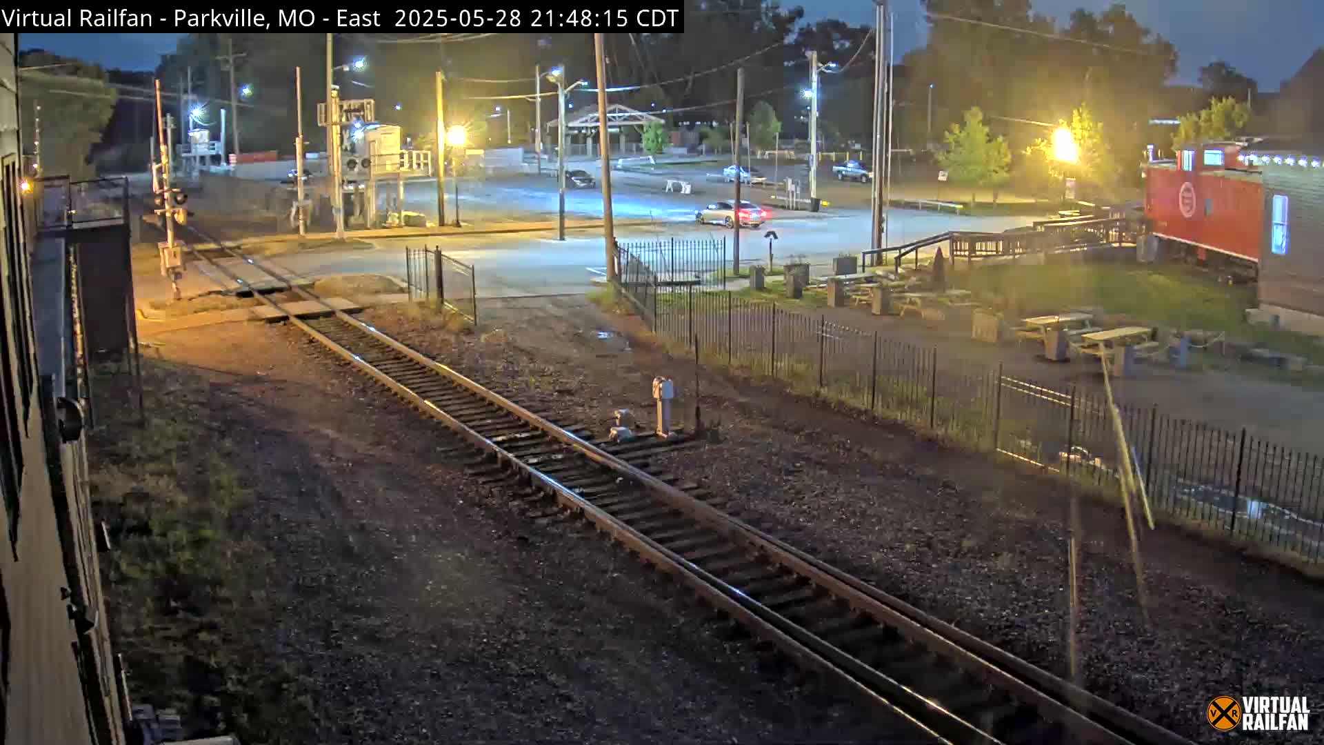 A nighttime view of railroad tracks intersecting a road, with a parking lot, some cars, and outdoor picnic tables visible in the background under artificial light.