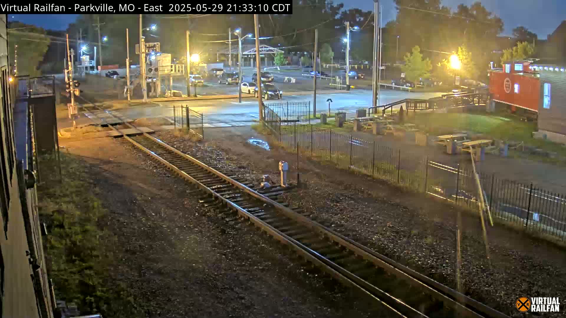 A nighttime view of railroad tracks intersecting a road, with a parking lot, some buildings, and outdoor seating visible under slightly wet conditions.