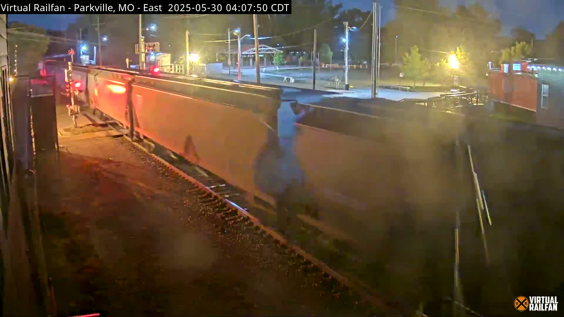 A long freight train passes by at night under slightly wet conditions, with a person visible on the side of one of the cars.
