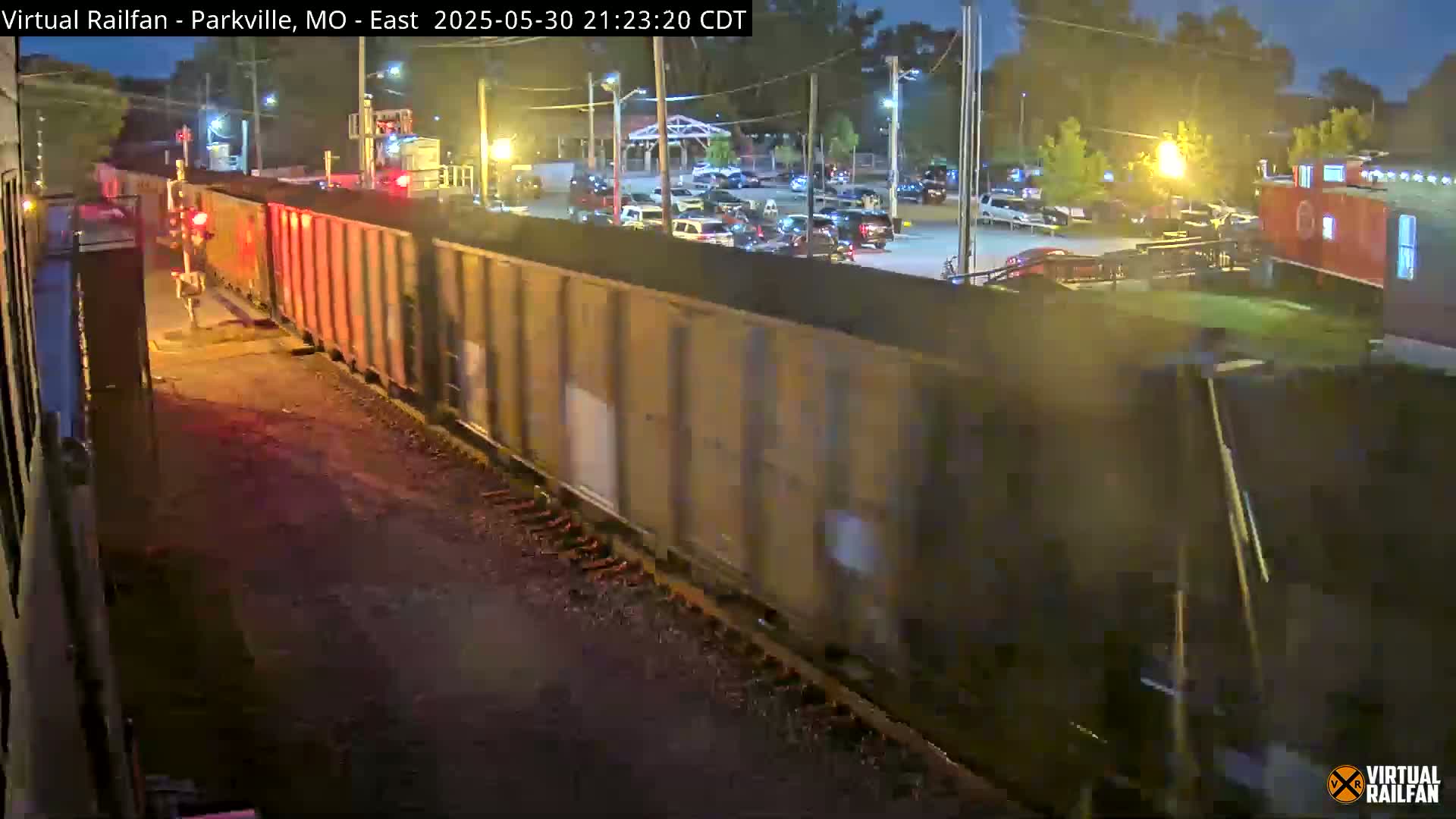 A long freight train passes at night near a parking lot with cars, under streetlights.