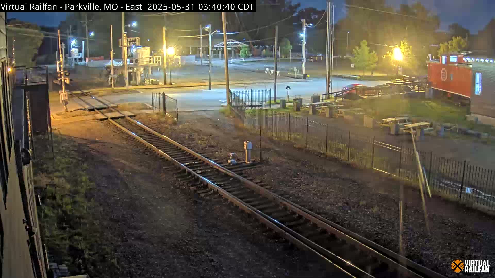 A nighttime view of railroad tracks intersecting a road, with a converted railcar structure and several outdoor picnic tables visible in the background under streetlights.
