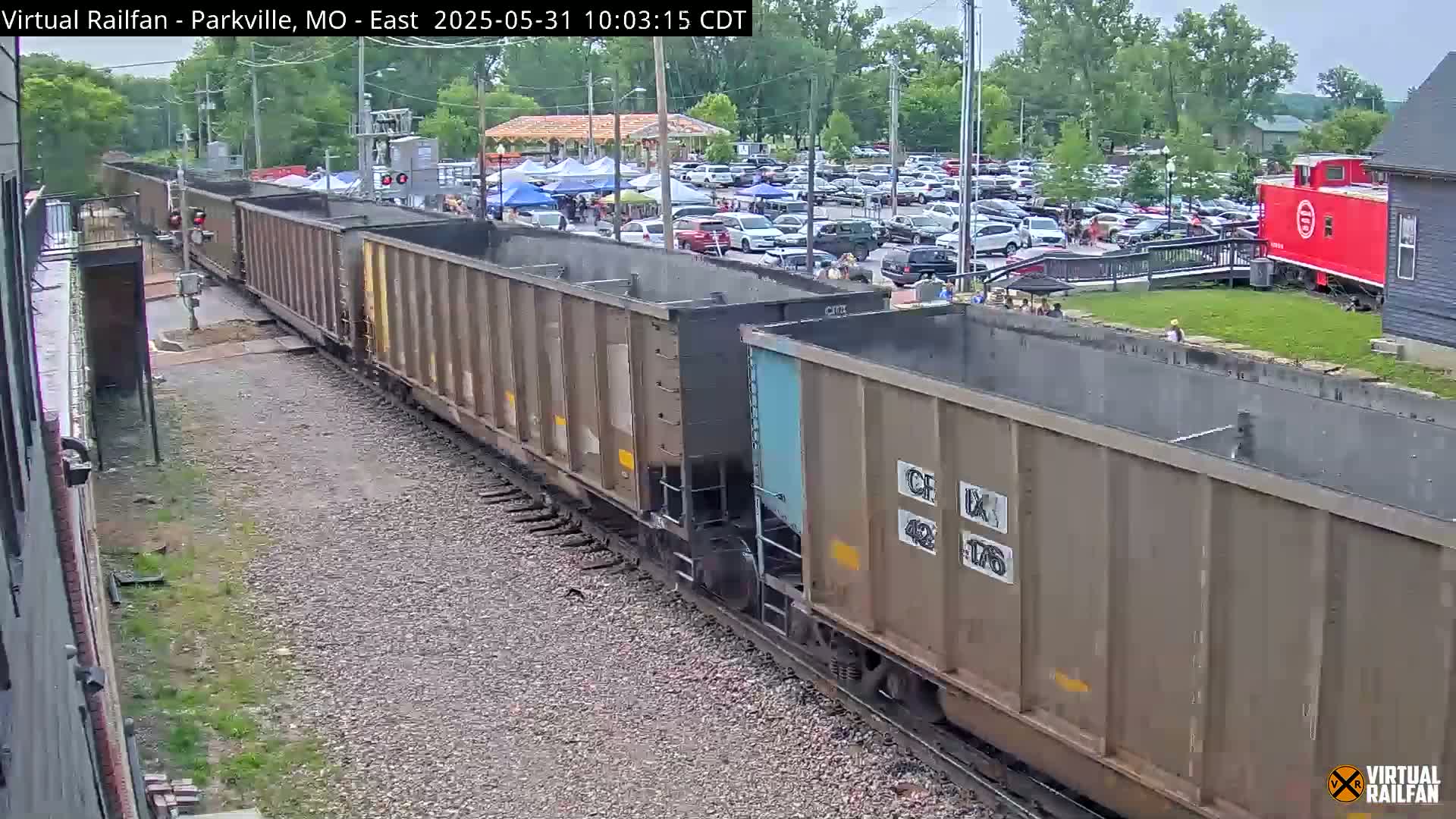 A long freight train of hopper cars passes by a parking lot filled with cars under an overcast sky.