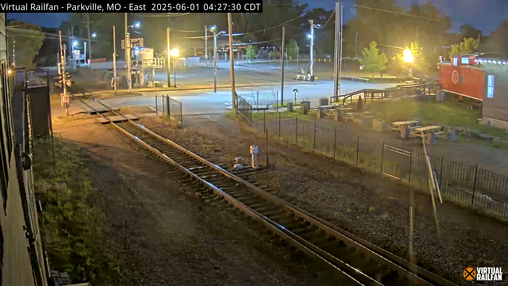 At night, train tracks cross a road near a building and outdoor seating area under illuminated streetlights.