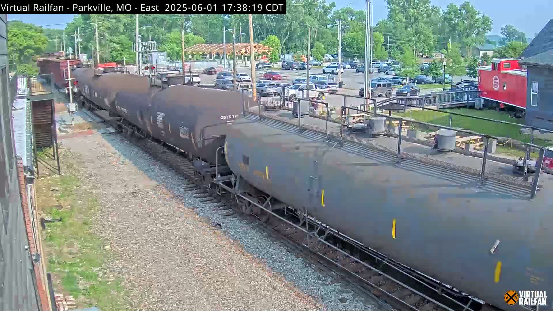 A long freight train of tanker cars moves along a railway track on a sunny day, passing by a parking lot and a red caboose near a building.