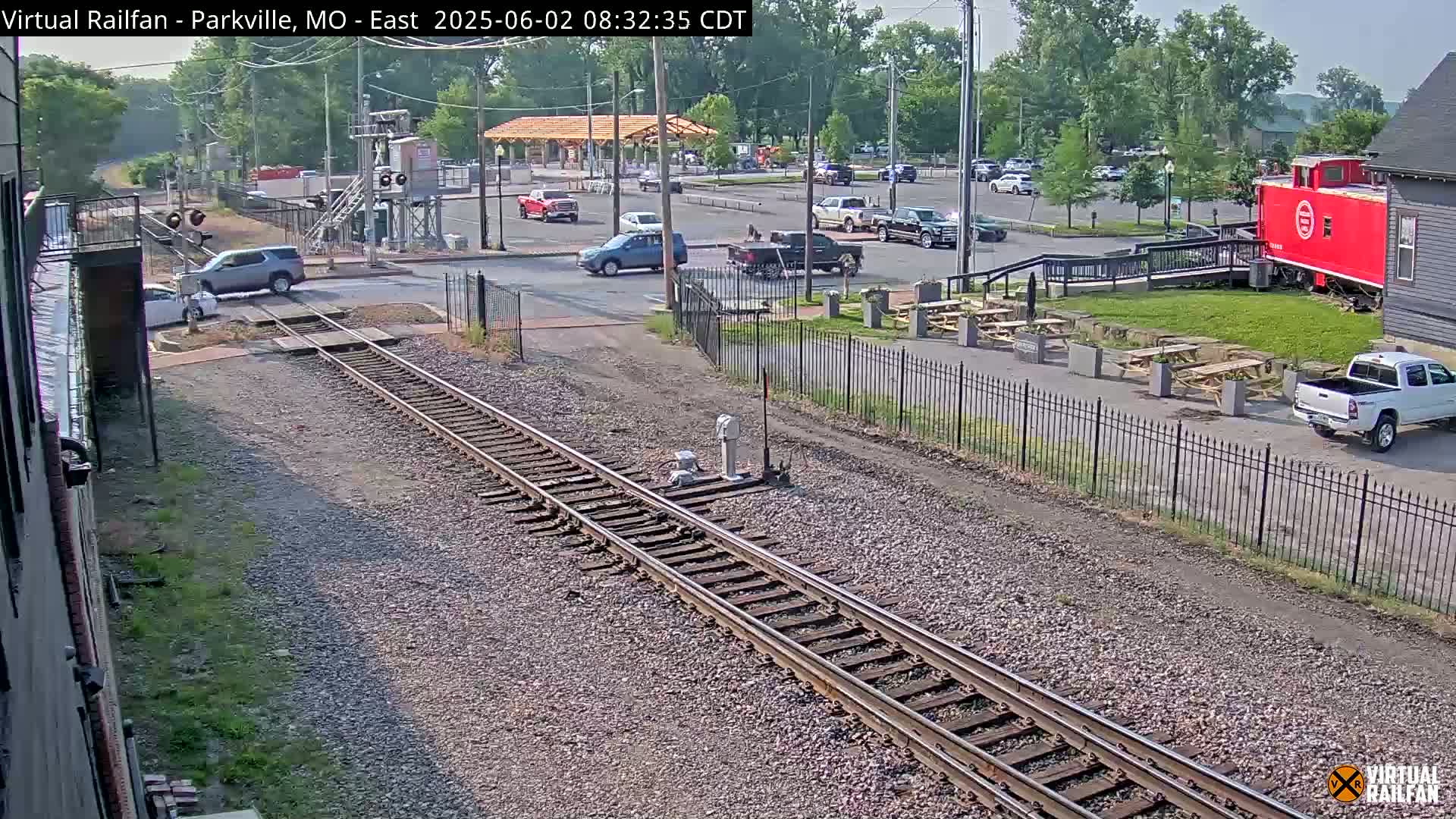 A sunny day view of railroad tracks intersecting a road with several vehicles, a red caboose, and a parking lot with picnic tables visible beyond a black metal fence.