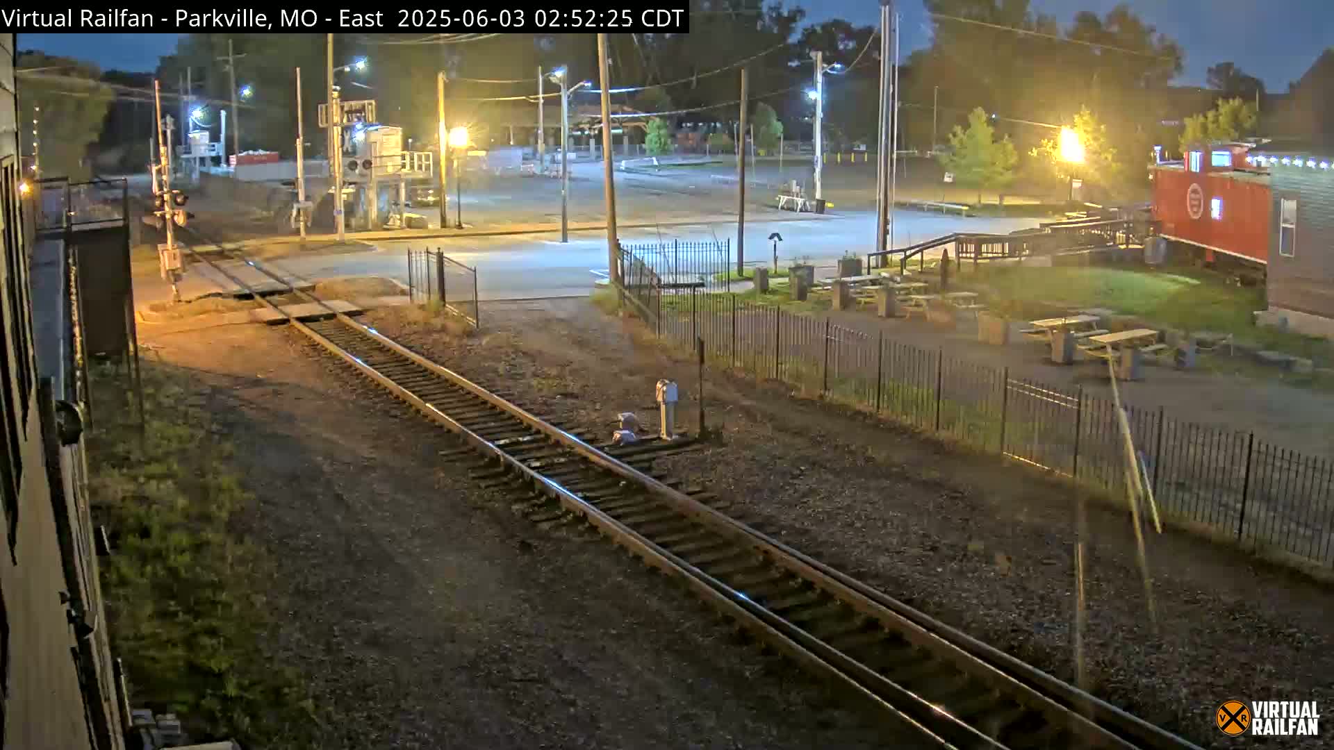 A nighttime view of railroad tracks intersecting a road, with a red caboose and picnic tables visible in the background under streetlights.