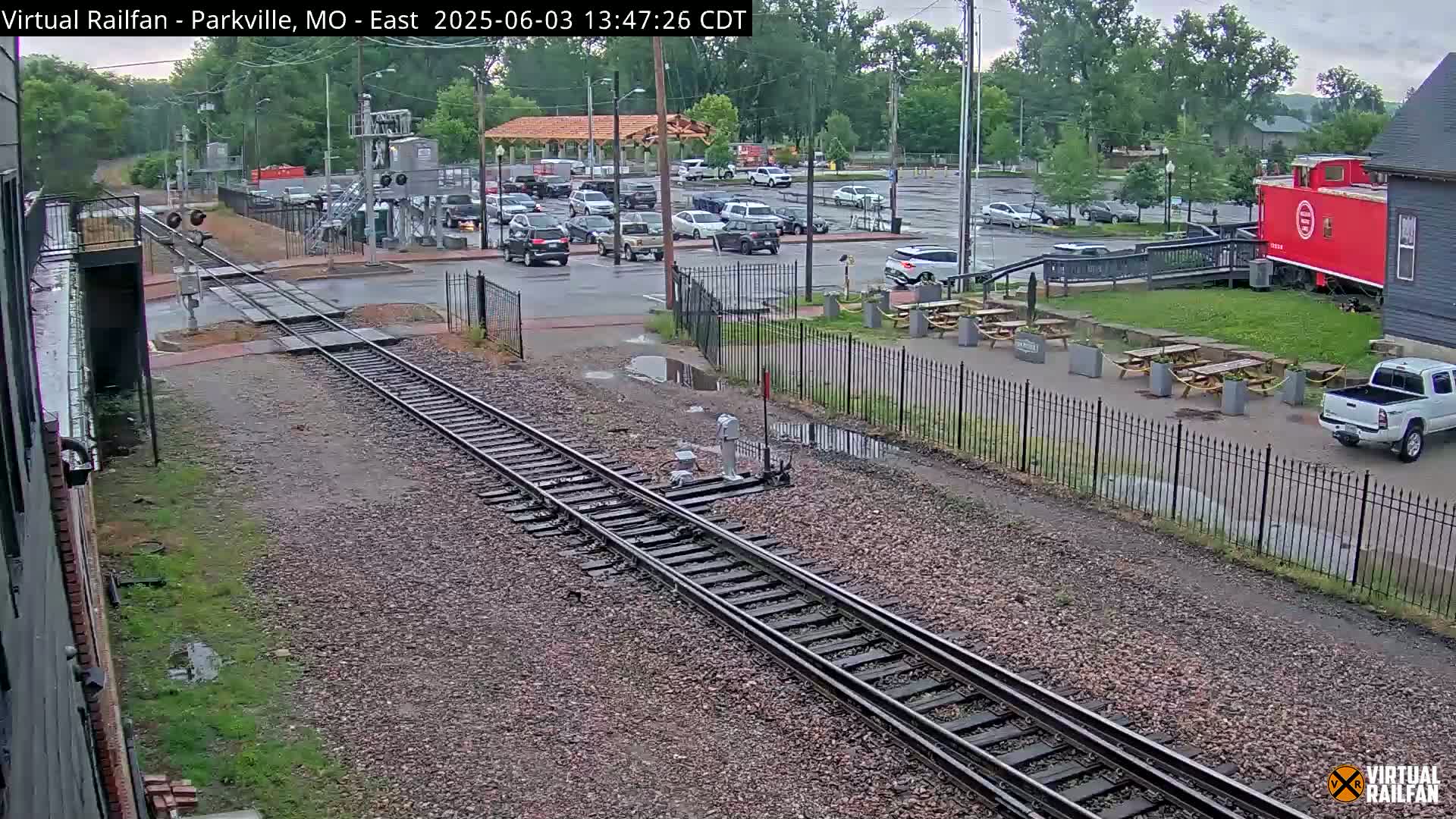 Railroad tracks cross a street with parked cars and a red caboose next to a building, under an overcast sky.