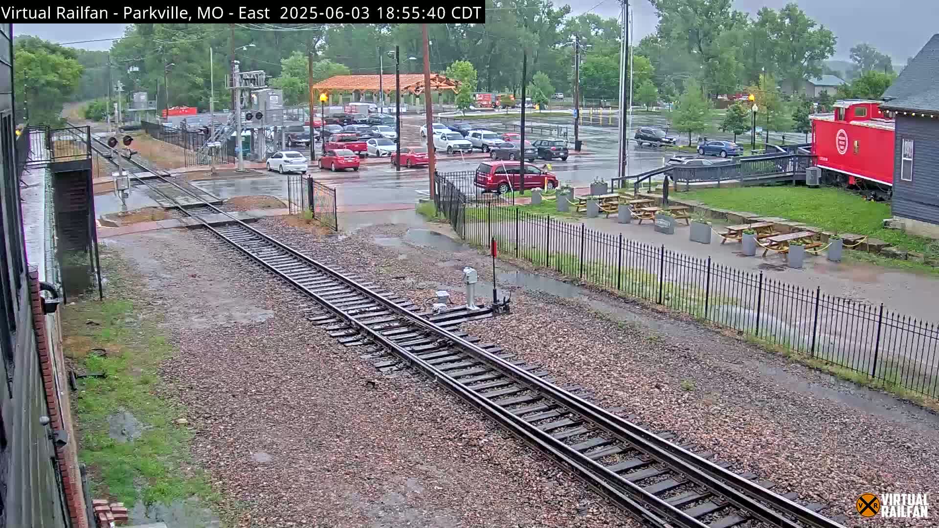 Rain falls on a railway crossing near a parking lot with several cars, a red caboose, and outdoor picnic tables.