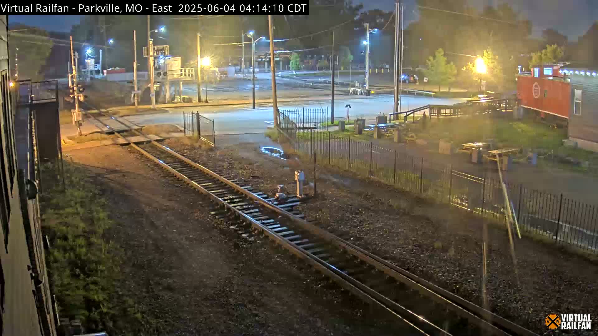 A nighttime view of railroad tracks intersecting a road, with a red caboose and other buildings visible in the background under wet conditions.