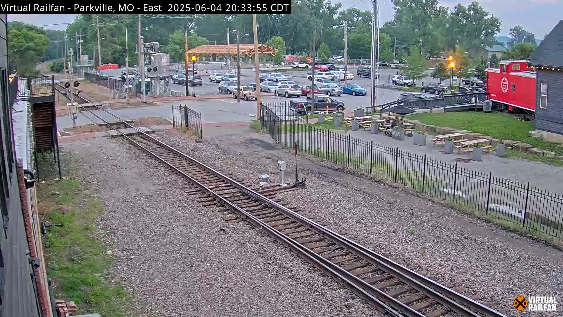 A railroad track runs through a parking lot with cars, a red caboose, and picnic tables under an overcast sky.