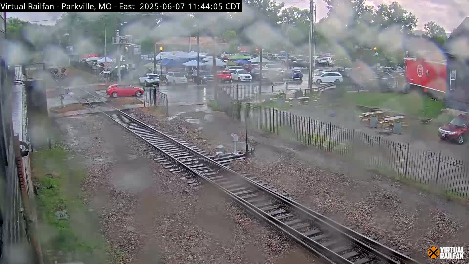 A rain-soaked parking lot and railroad crossing are seen from a train's perspective, with several cars parked nearby and a few people walking.