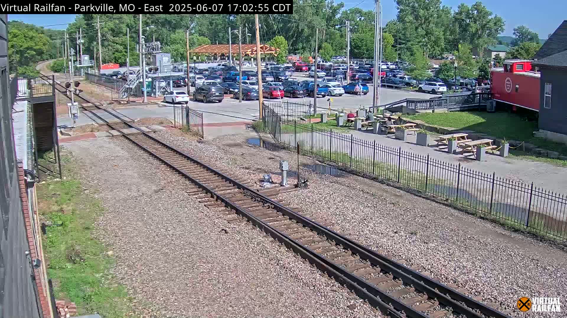 Railroad tracks in a sunny outdoor setting pass by a parking lot full of cars, a building with outdoor picnic tables, and a red caboose.