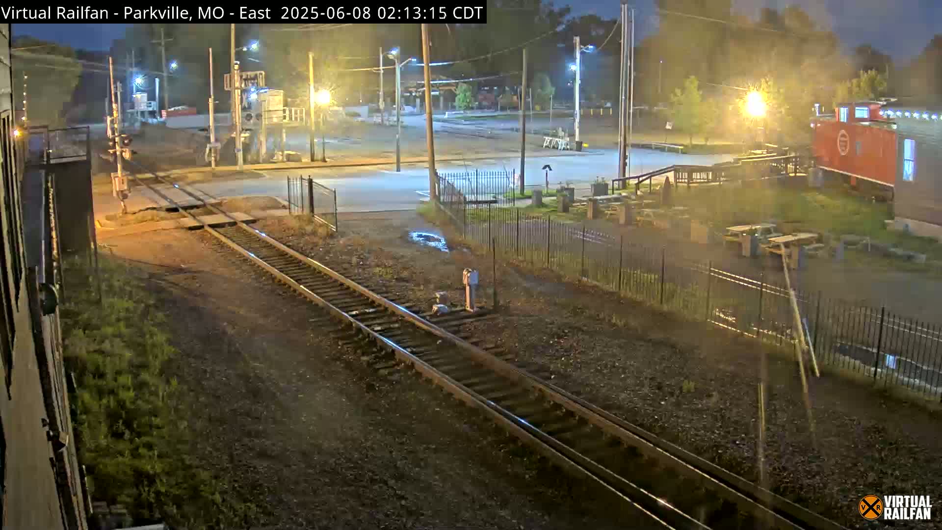 A nighttime view of railroad tracks intersecting a road, with a red caboose-like structure visible in the background under partly cloudy skies.