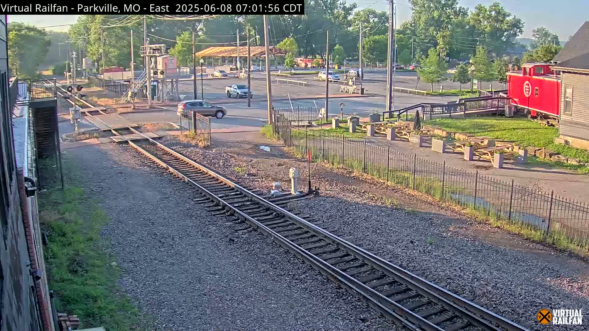 A sunny view of railroad tracks intersecting with a road near a red caboose and a parking lot.