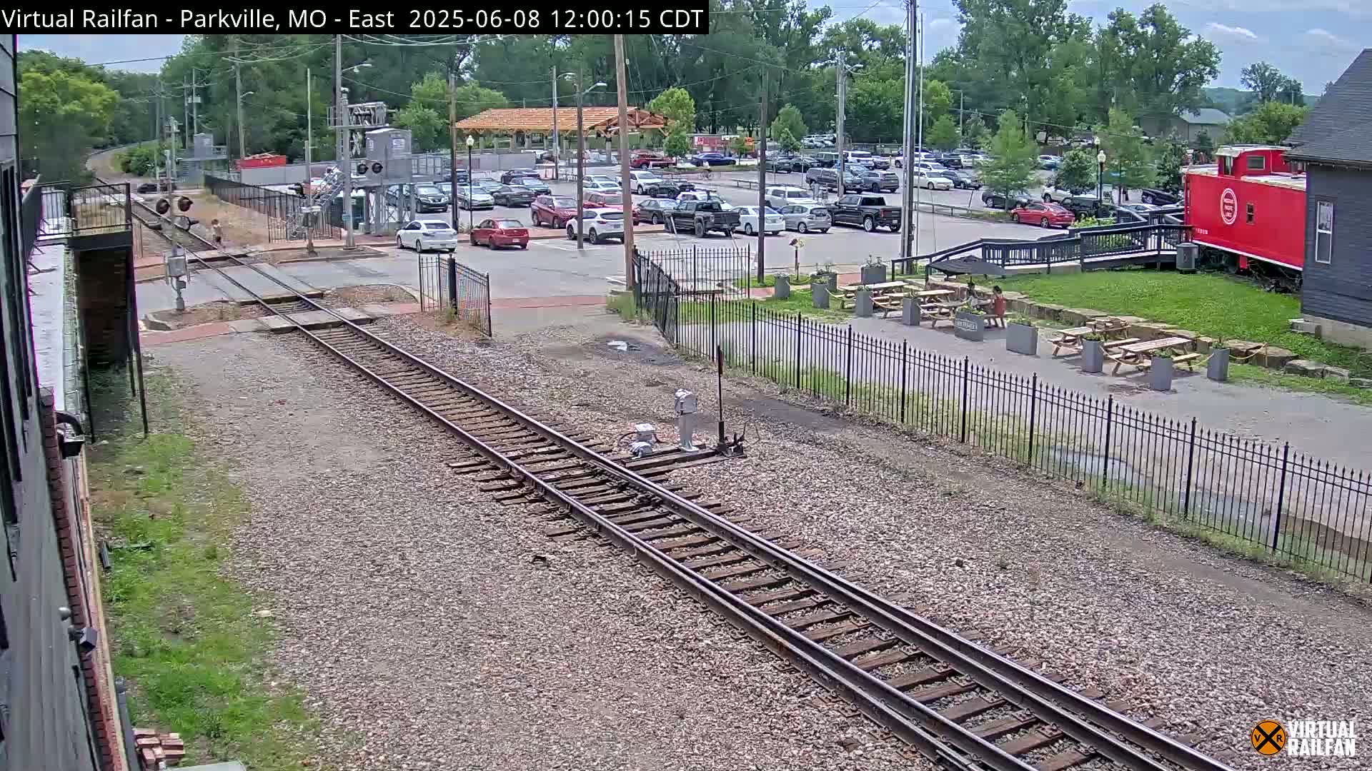 A view of railroad tracks intersecting a roadway next to a parking lot and outdoor seating area on a partly sunny day.