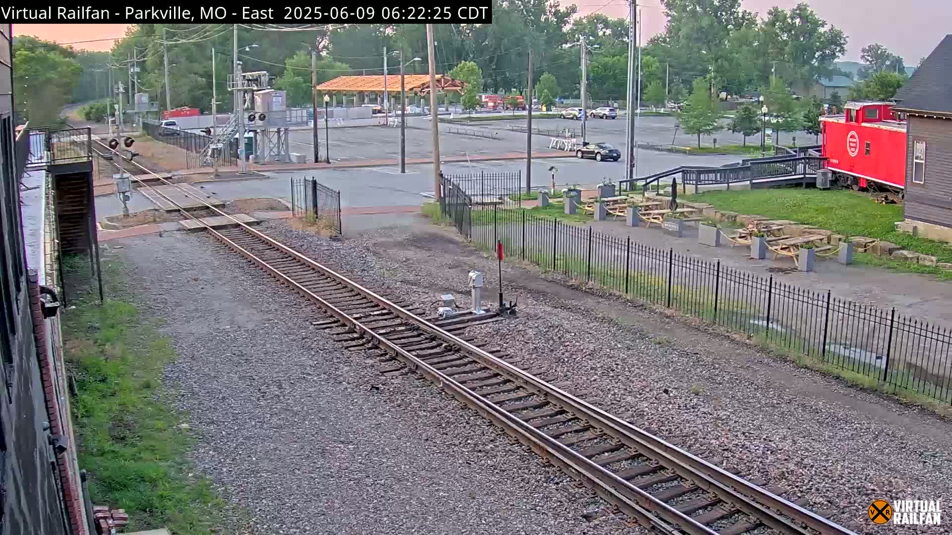 Railroad tracks extend from the foreground toward a parking lot with a red caboose and picnic tables under a partly cloudy sky.