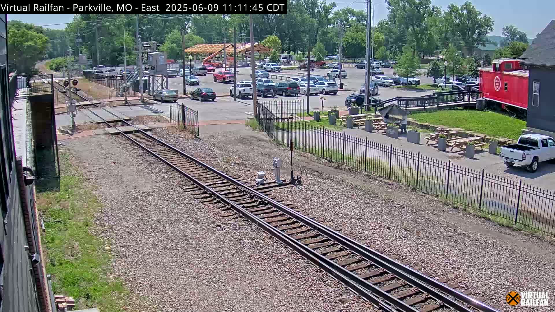 A railroad crossing with train tracks in the foreground, a parking lot with numerous vehicles, an outdoor seating area with picnic tables, and a red caboose visible in the background on a sunny day.