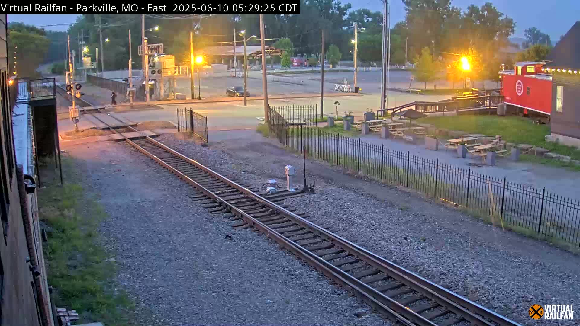 A twilight view of railroad tracks intersecting a road near a building with picnic tables and a red caboose.