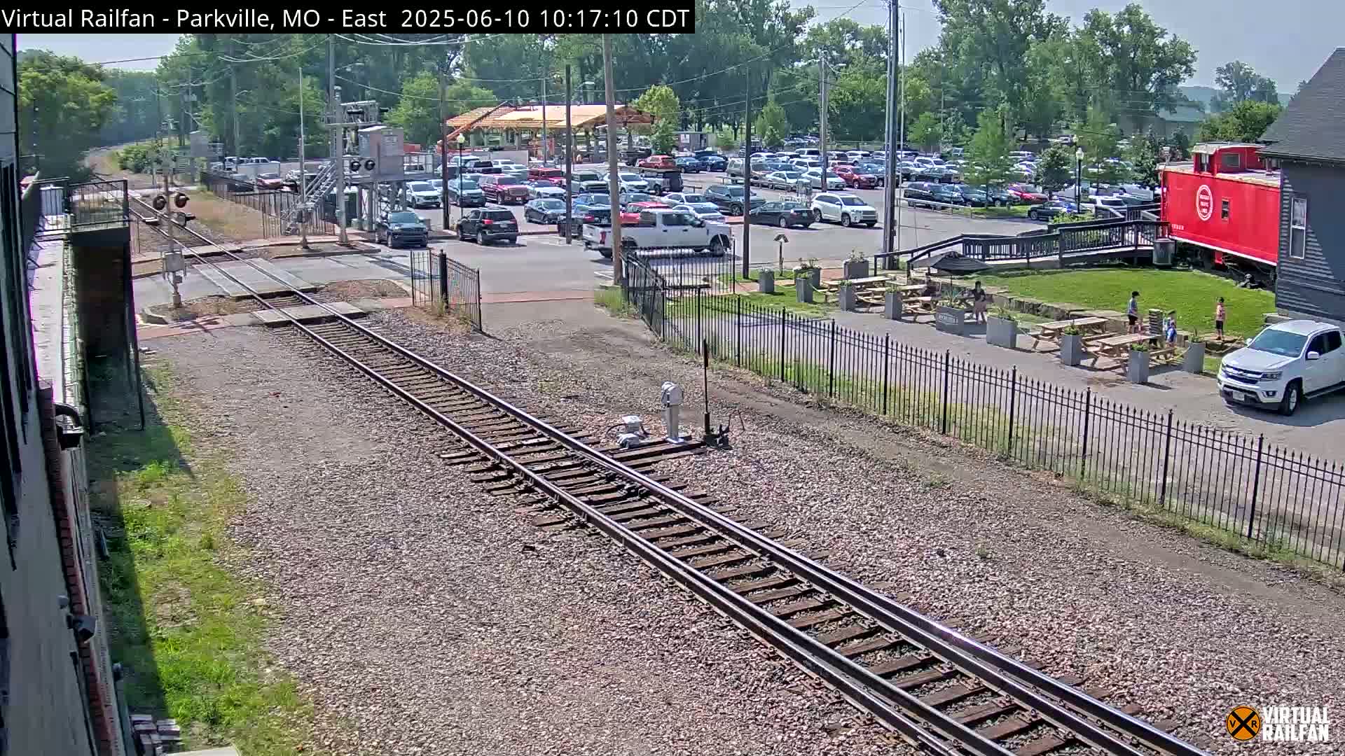 A sunny view of railroad tracks next to a parking lot and outdoor seating area, with a red caboose visible in the background.