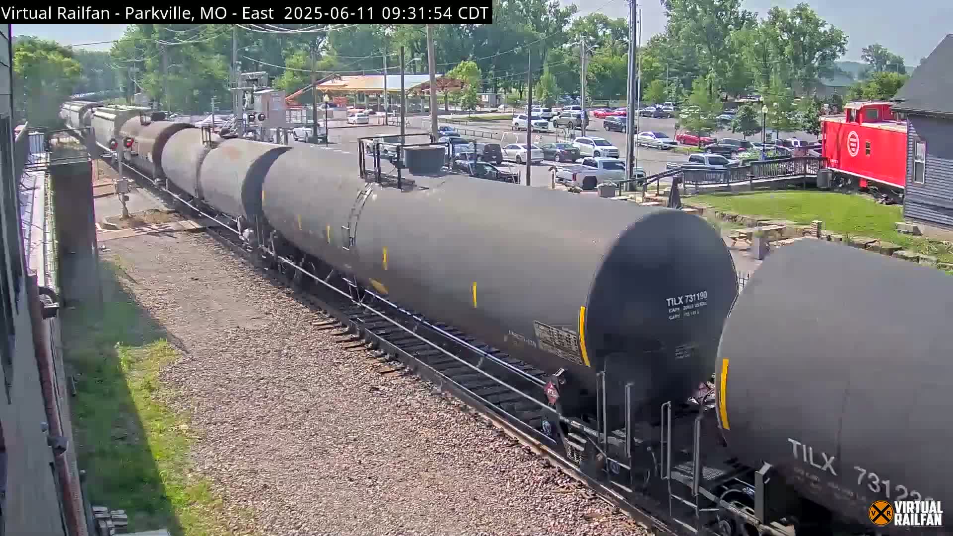 A long train of tanker cars moves along railway tracks on a sunny day, past a parking lot with many cars and a red caboose.