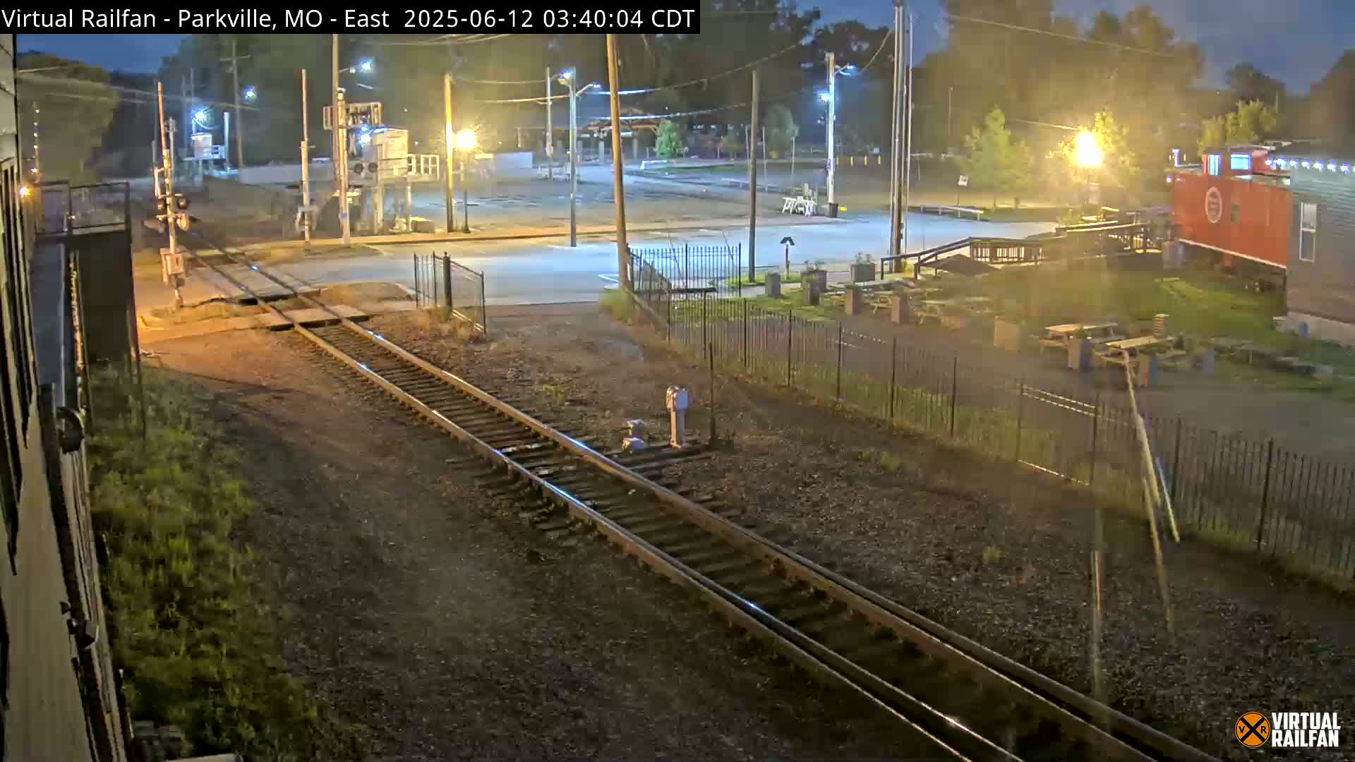 At night, railroad tracks intersect with a road next to a fenced-in area with picnic tables and a red caboose.