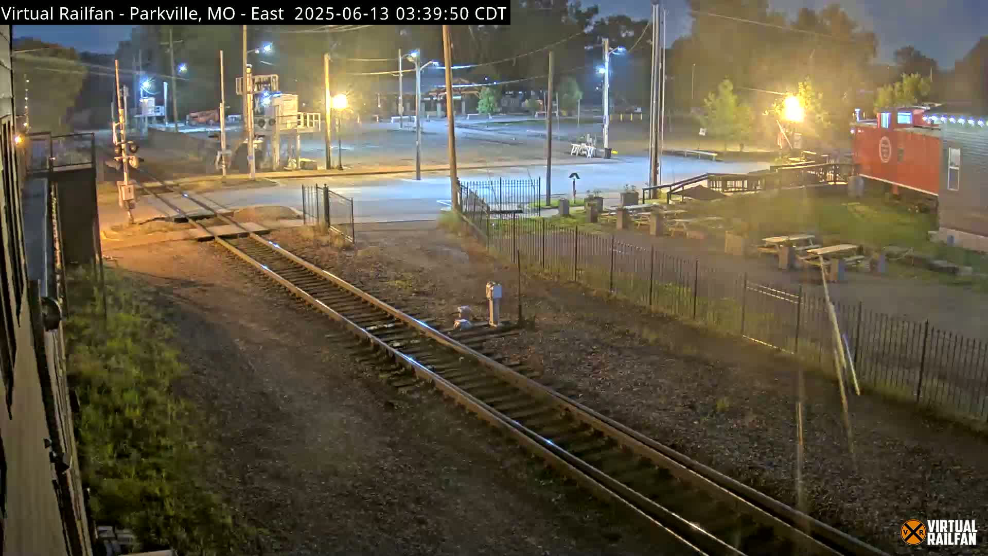 A nighttime view of railroad tracks intersecting with a road, showing a red caboose and some outdoor seating in the background under artificial light.