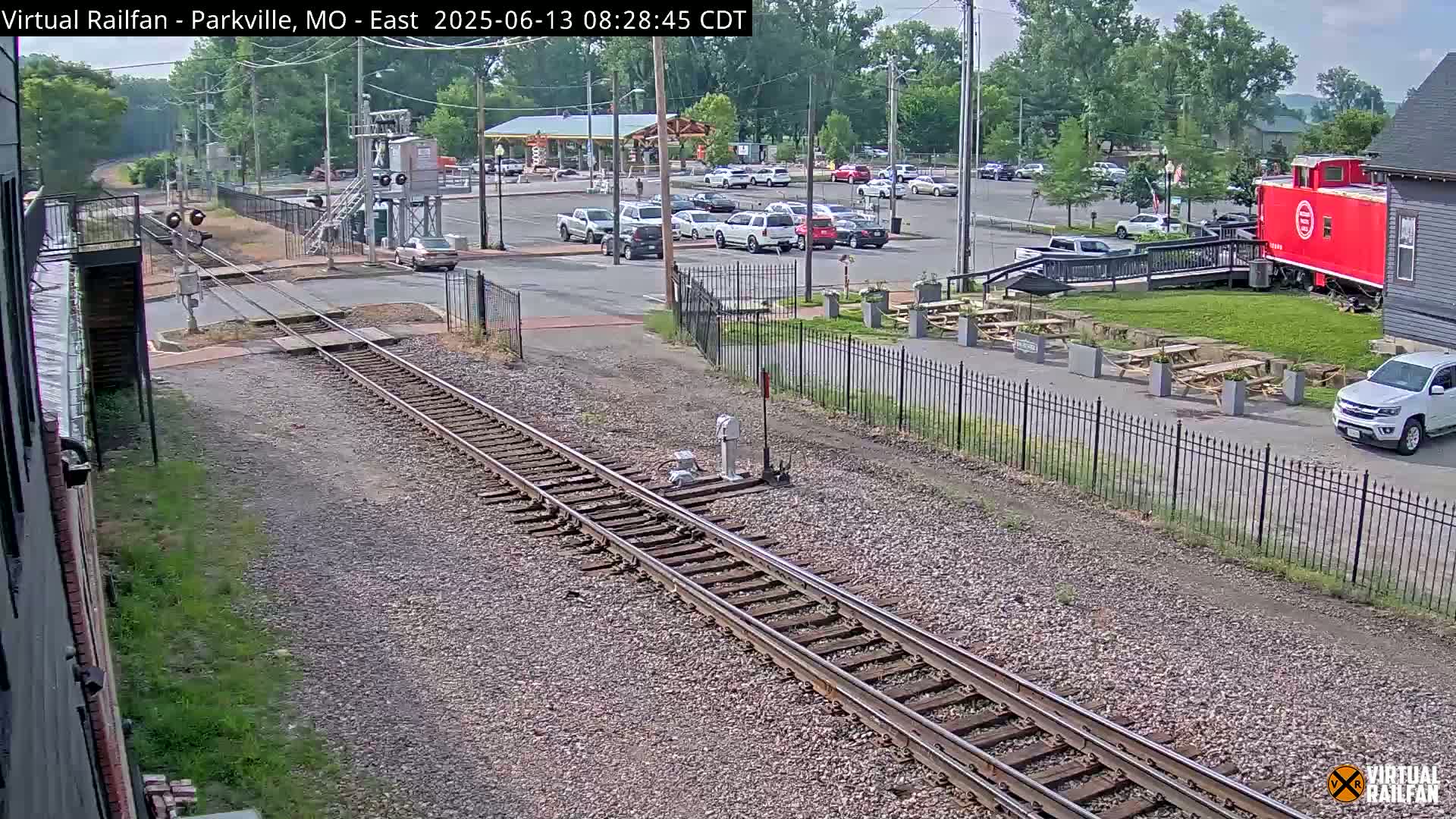 A view of railroad tracks on a sunny day, with a parking lot, a red caboose, and several picnic tables visible in the background.