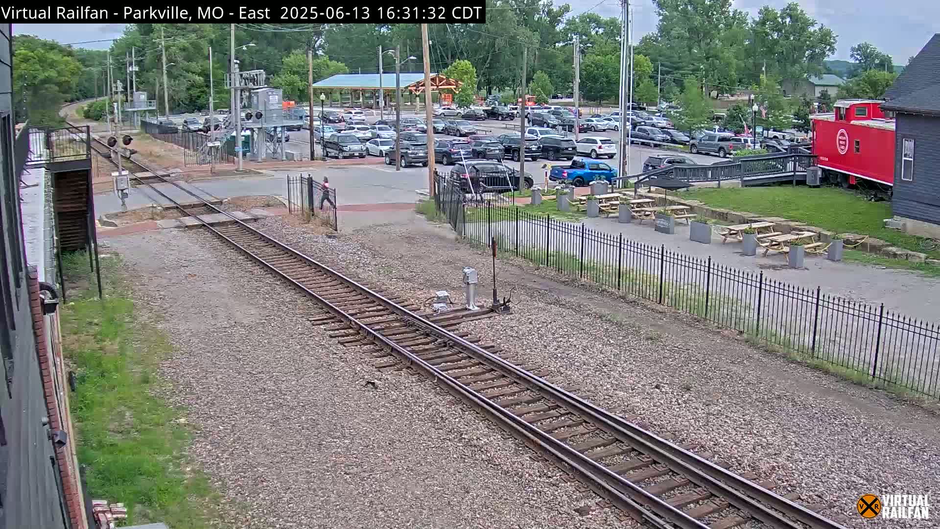 A mostly sunny day reveals railroad tracks crossing a road near a parking lot with several cars, a red caboose, and picnic tables.