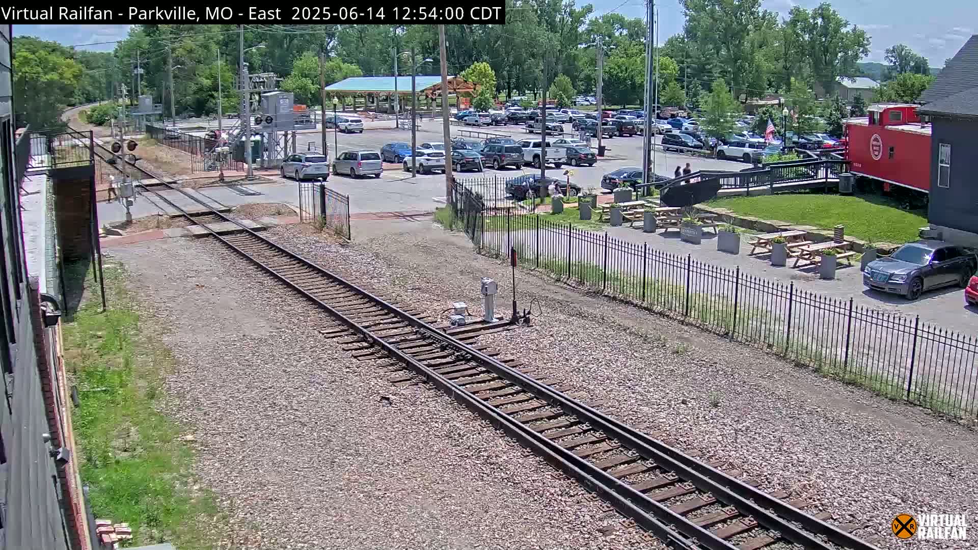 A sunny day shows a railroad crossing with train tracks, a parking lot full of cars, and a red caboose next to a building with picnic tables outside.