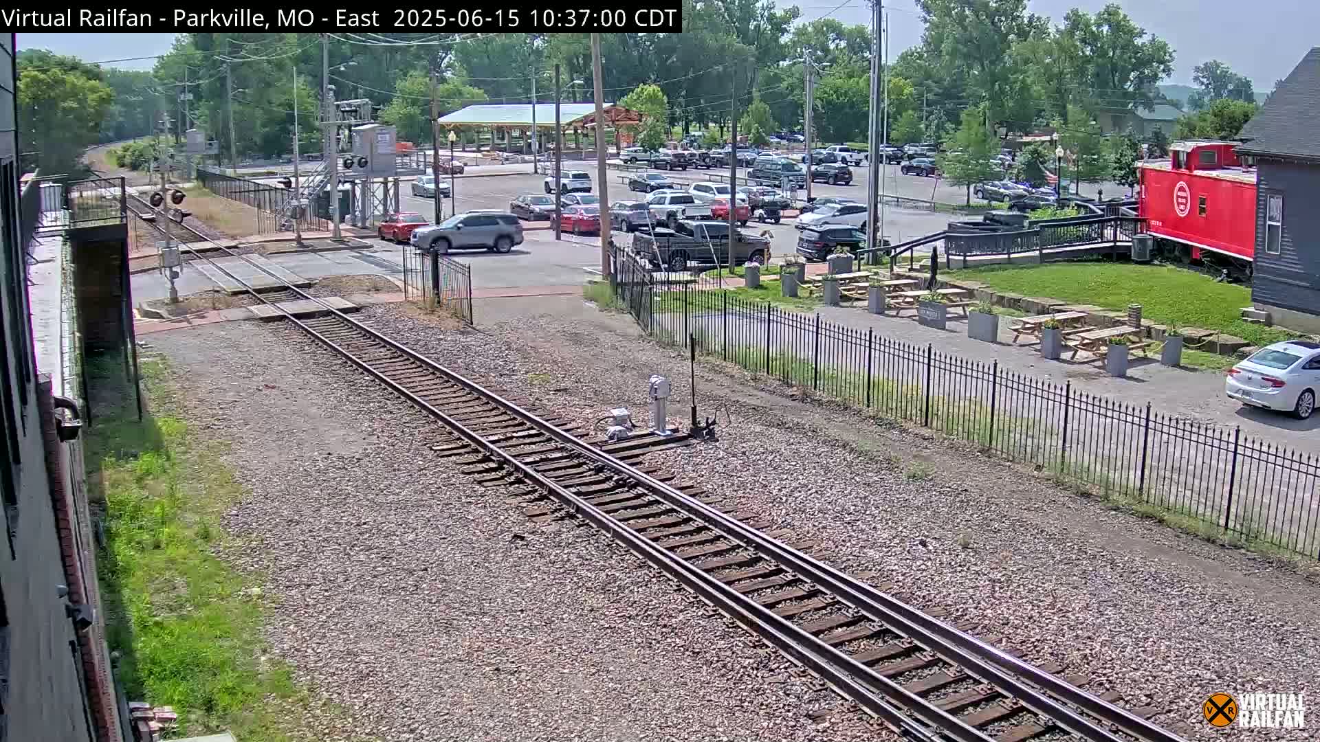A sunny day shows railroad tracks extending from a parking lot with cars and a red caboose near a building, all enclosed by a black metal fence.