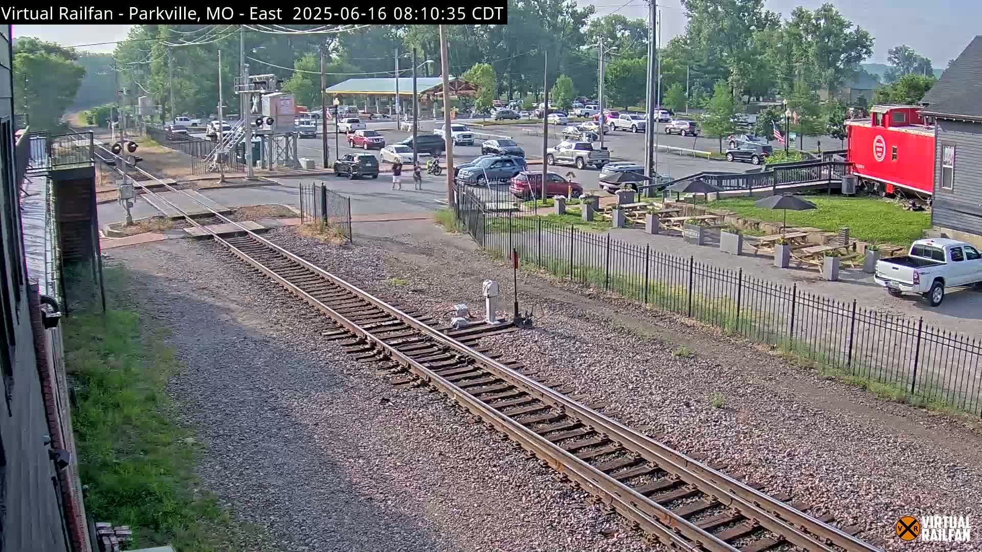 Railroad tracks intersect a street with parked cars, a small building, and outdoor seating under a sunny sky.