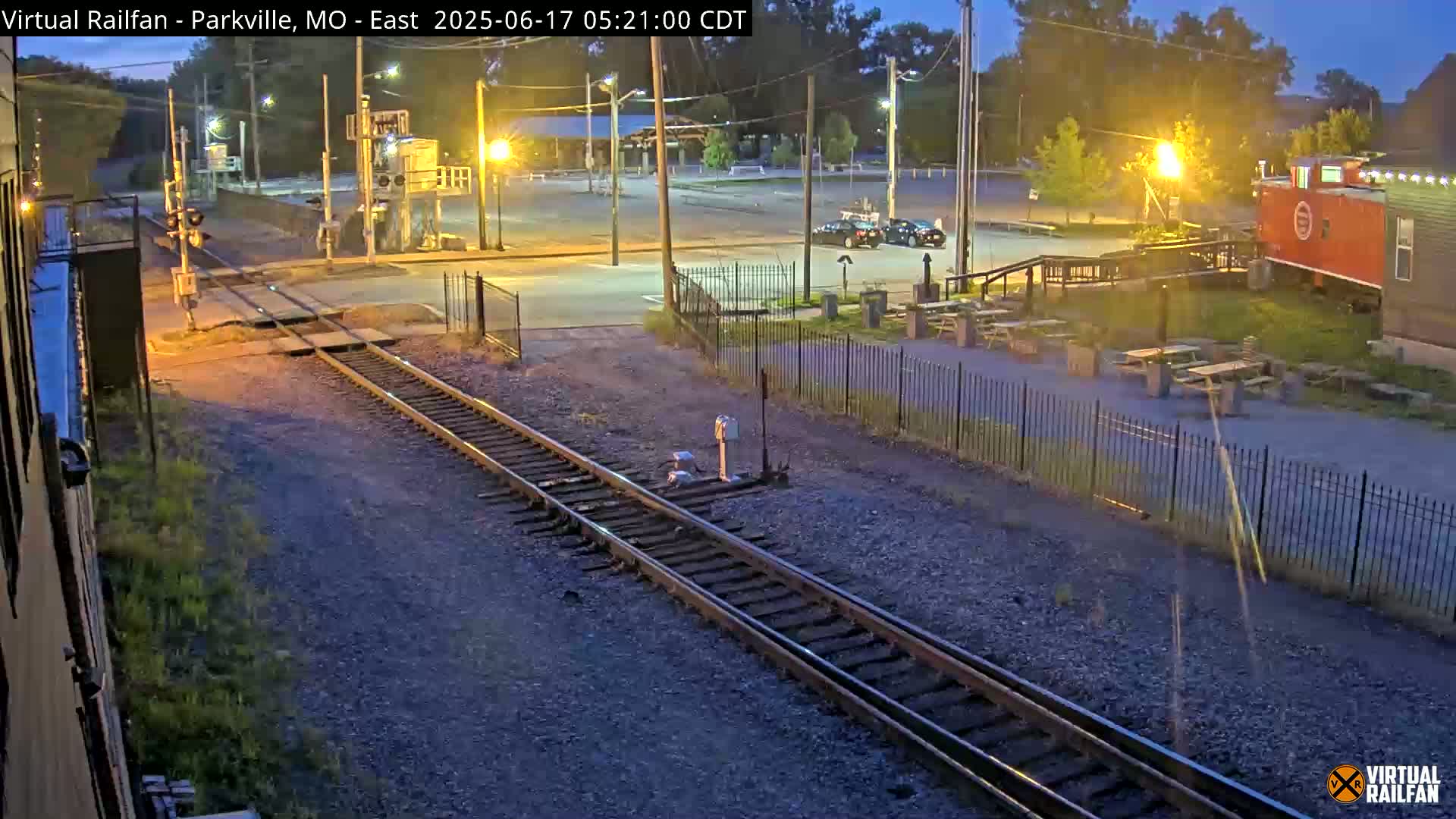 Railroad tracks intersect a road at dusk, next to a parking lot with a few cars, and a red caboose sits beyond a fence near some picnic tables.
