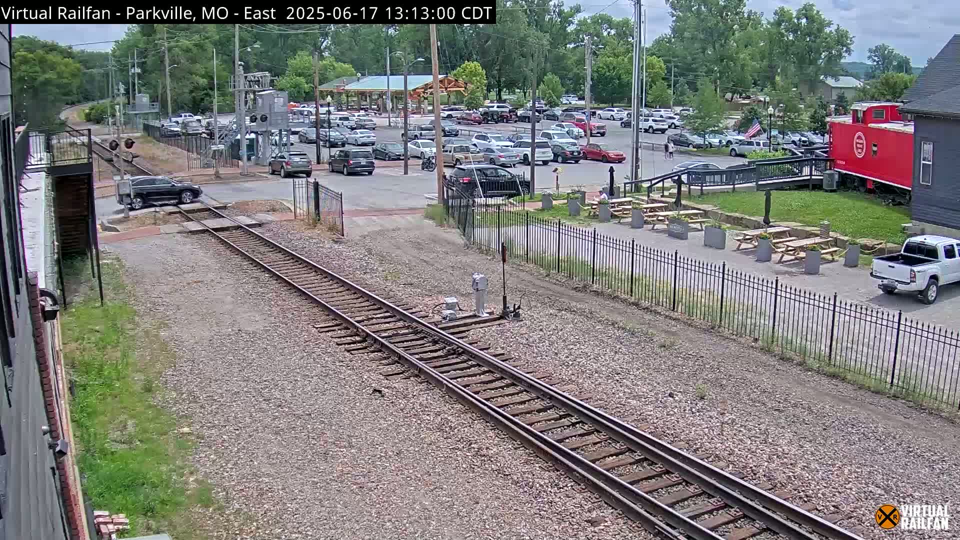 A dark-colored SUV crosses a railway track in a sunny outdoor setting, with a parking lot full of cars and a red caboose visible in the background.