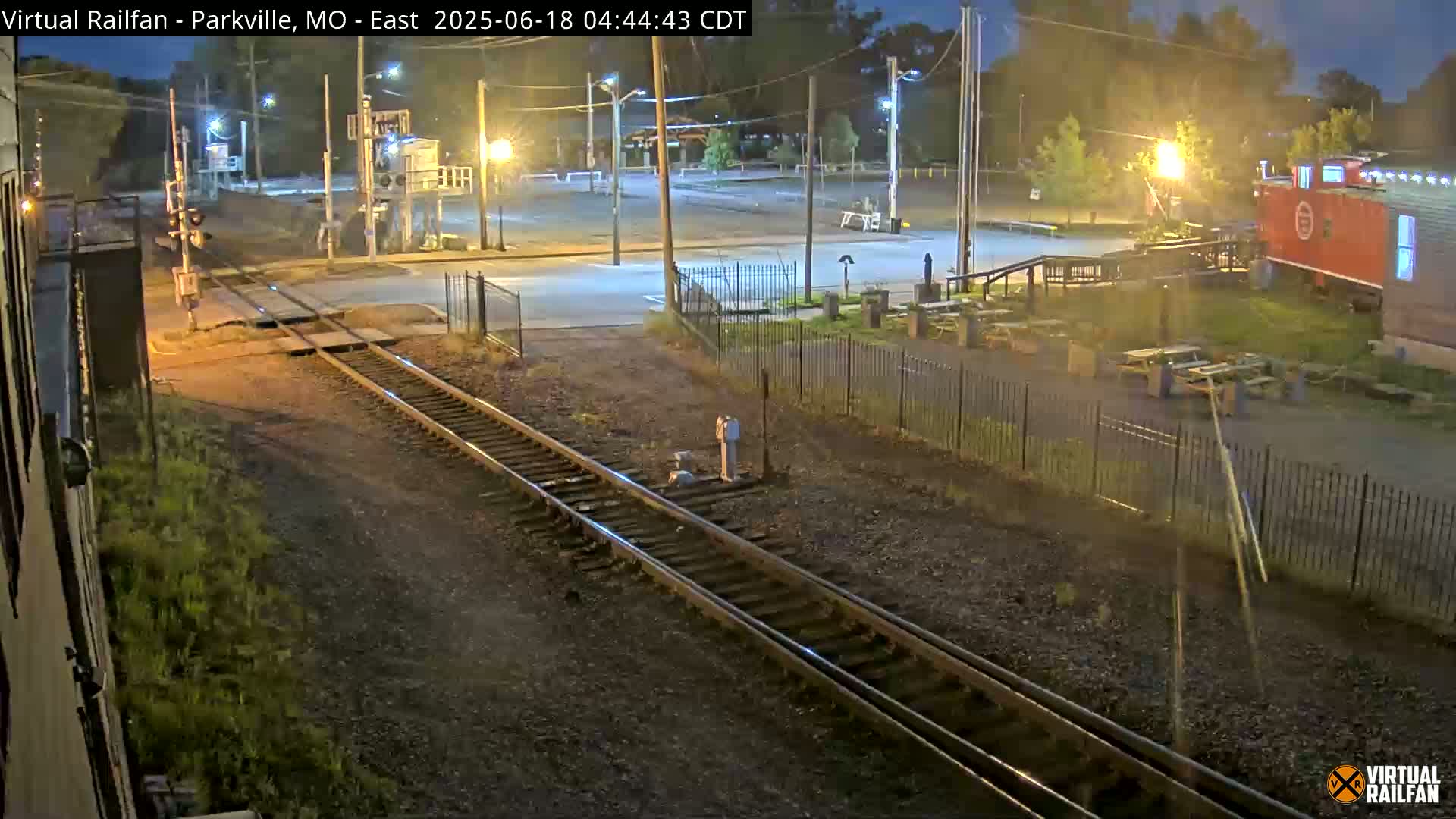 A nighttime view of railroad tracks intersecting a road, with a red caboose and a parking lot visible in the background under partly cloudy skies.