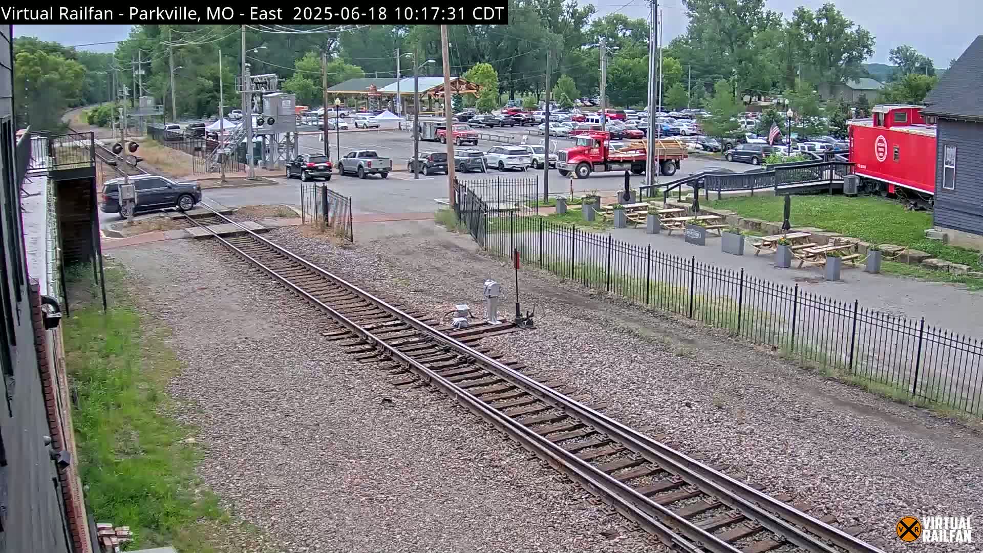 A view of railroad tracks leading to a parking lot with several vehicles and a red caboose under a partly cloudy sky.