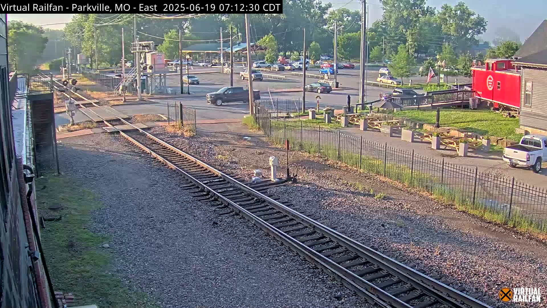 A mostly sunny outdoor scene shows railroad tracks intersecting a roadway with several vehicles parked and driving in a parking lot, a red caboose, and picnic tables by a building.