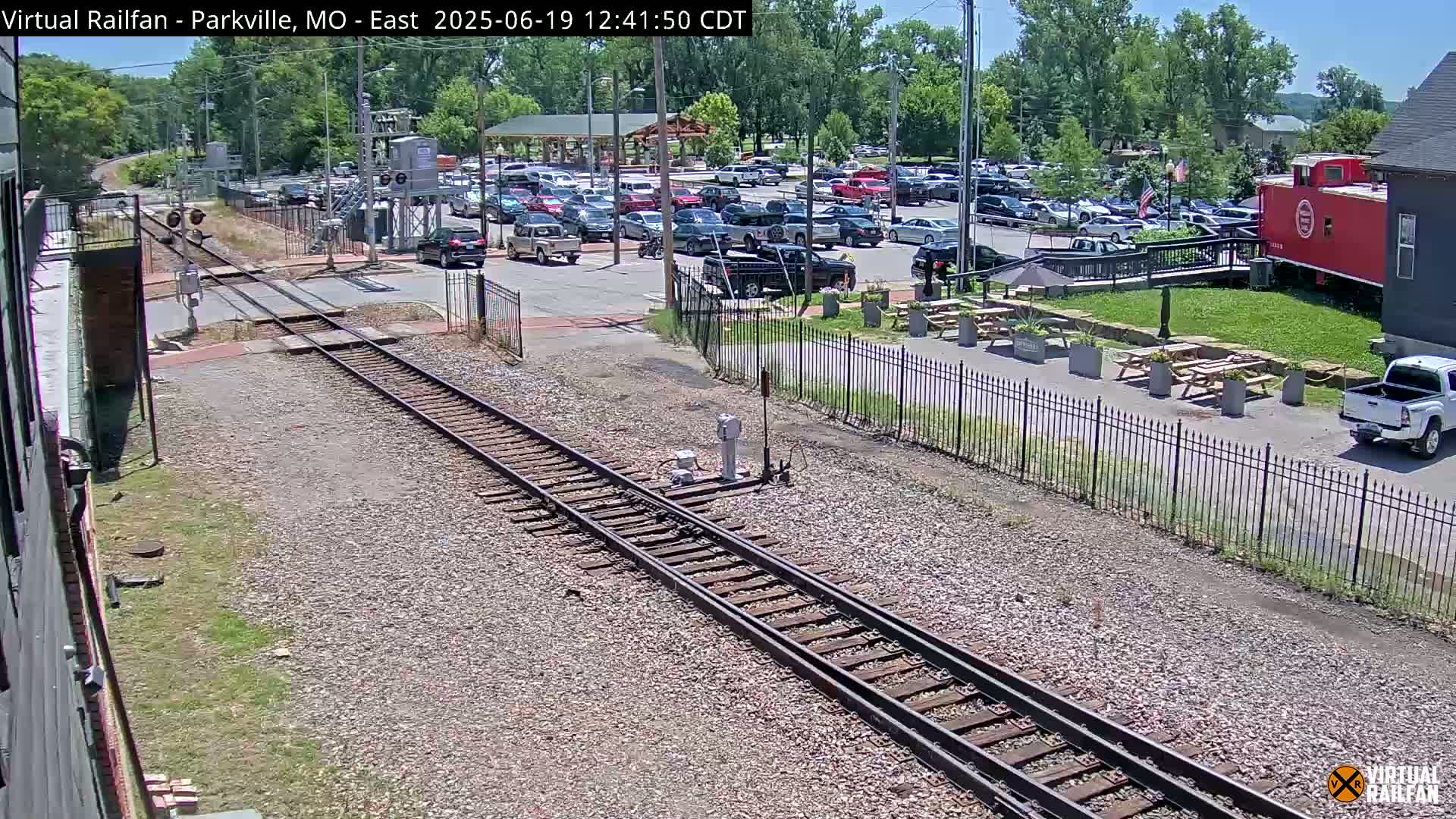 A sunny day view of railroad tracks intersecting with a roadway, beside a parking lot and a red caboose.
