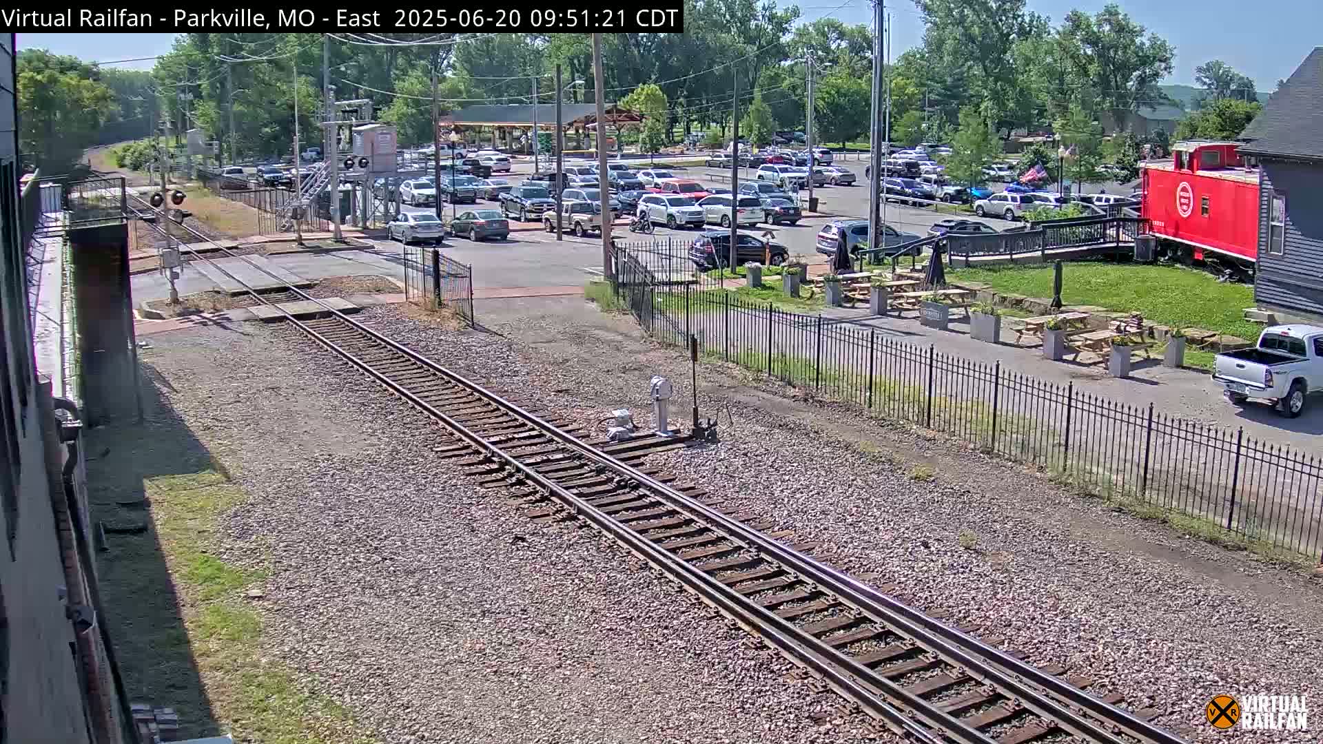 A sunny day reveals a railway crossing a parking lot filled with cars, near a red caboose and outdoor seating area.
