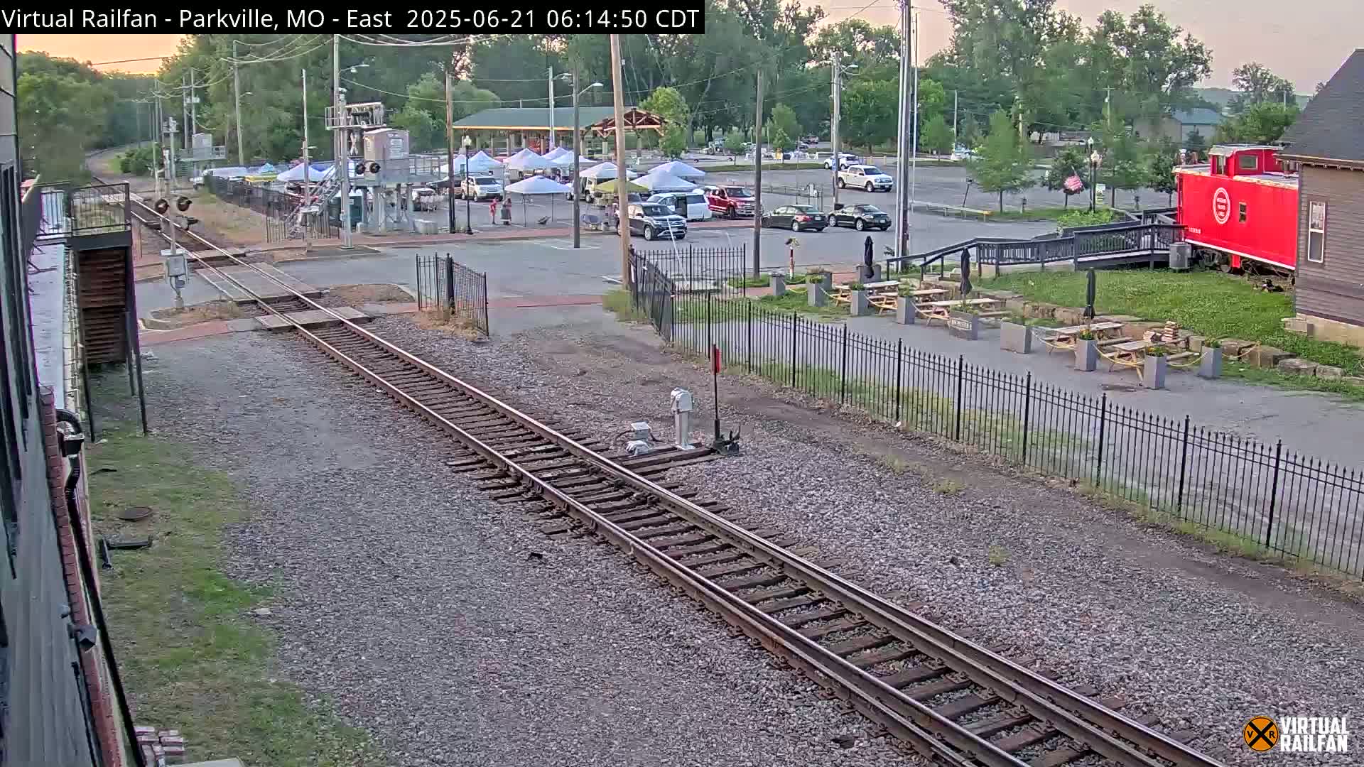 A railroad track runs through a parking lot with several cars and a red caboose next to an outdoor seating area under a partly cloudy evening sky.
