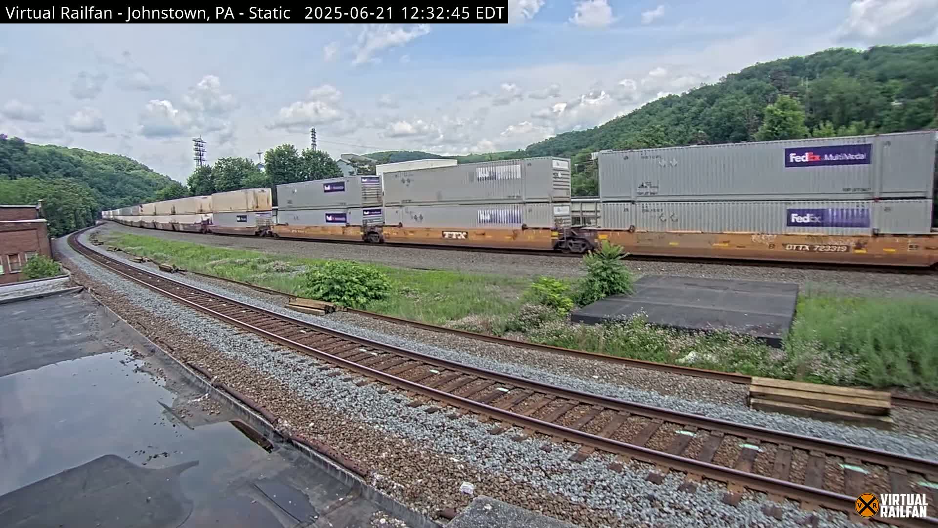 A long freight train carrying multiple shipping containers moves along railroad tracks under a partly cloudy sky.
