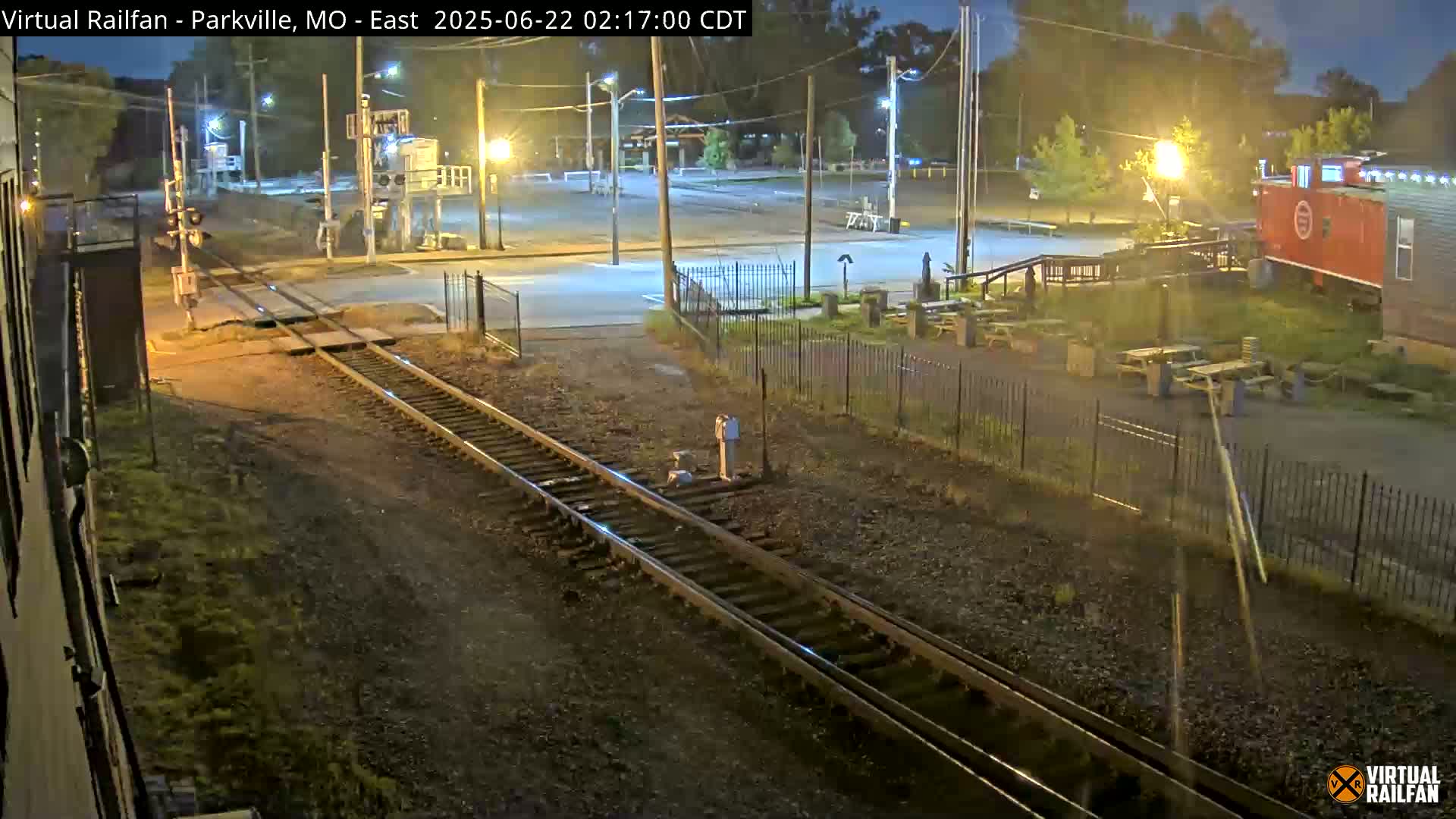 A nighttime view of railroad tracks intersecting a road, with a red caboose visible in the background under artificial light.