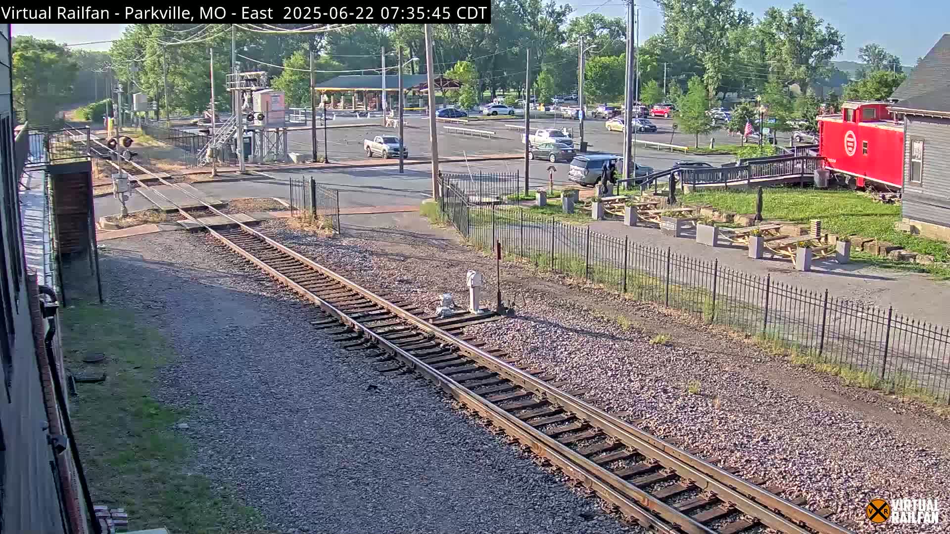 A sunny view of railroad tracks, a parking lot with cars, a red caboose, and picnic tables.