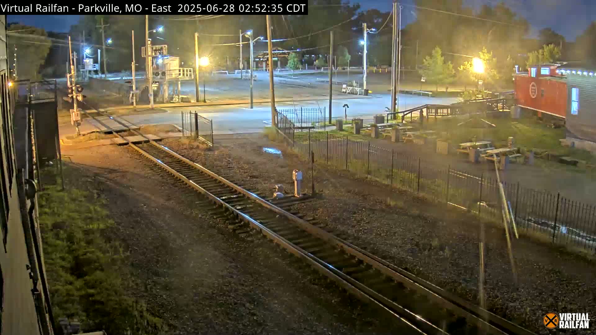 A nighttime view of railroad tracks intersecting a road, with a red caboose visible in the background under slightly wet conditions.