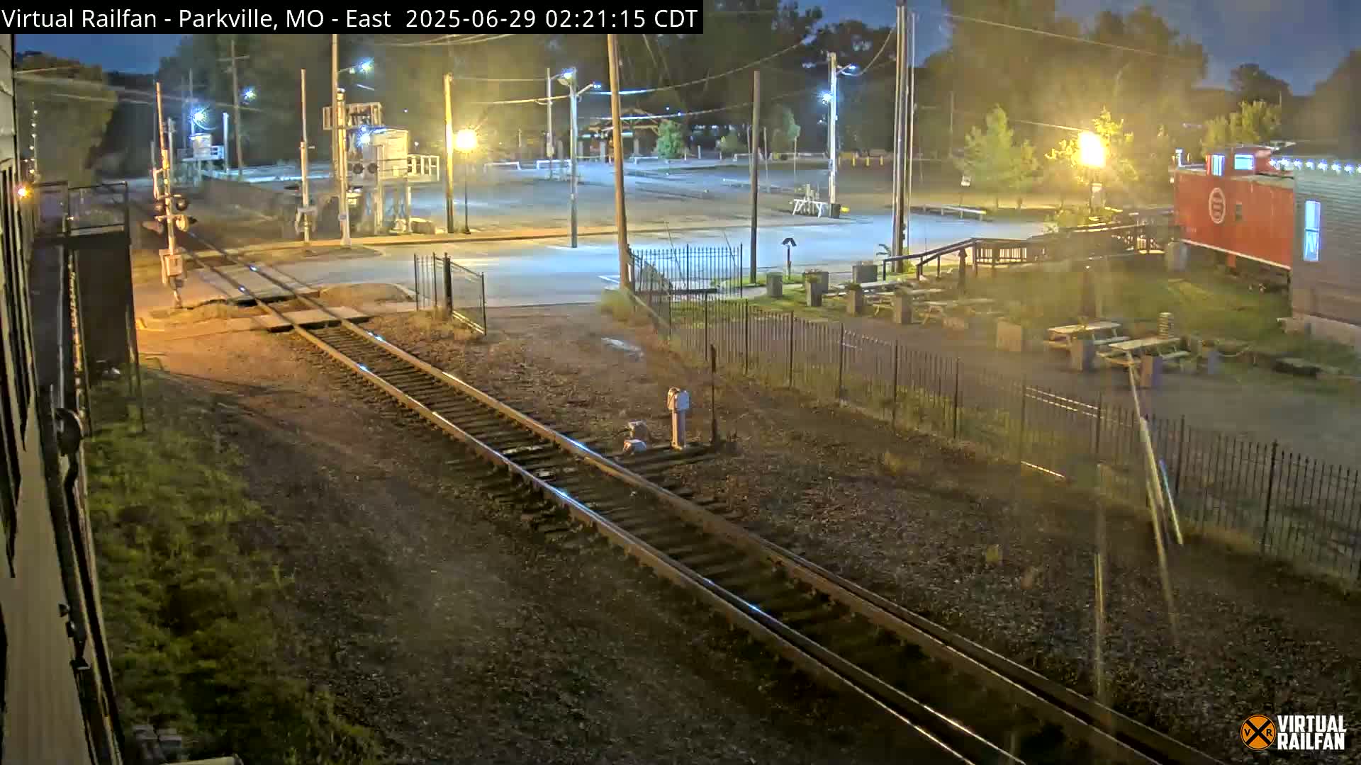 A nighttime view of railroad tracks intersecting a road, with a red caboose visible in the background under illuminated streetlights.