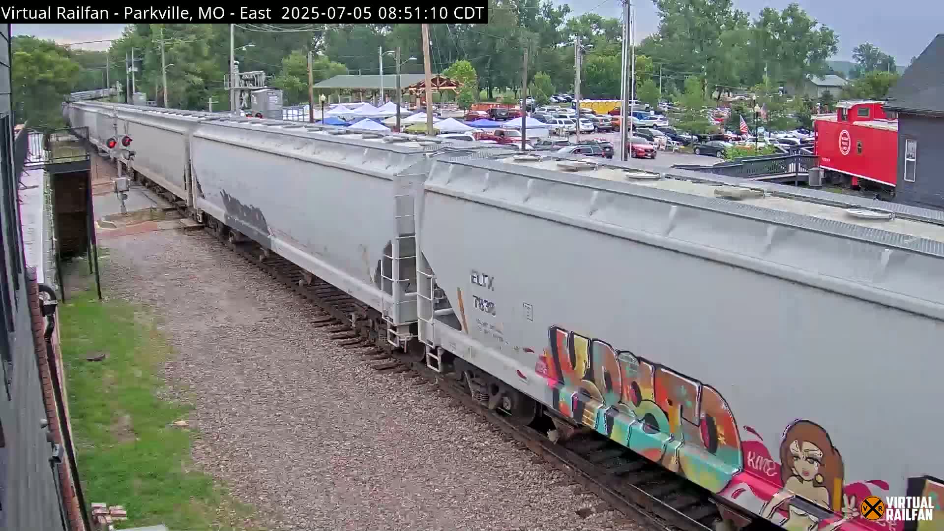 A long freight train with graffiti on its cars passes by a parking lot with cars under an overcast sky.