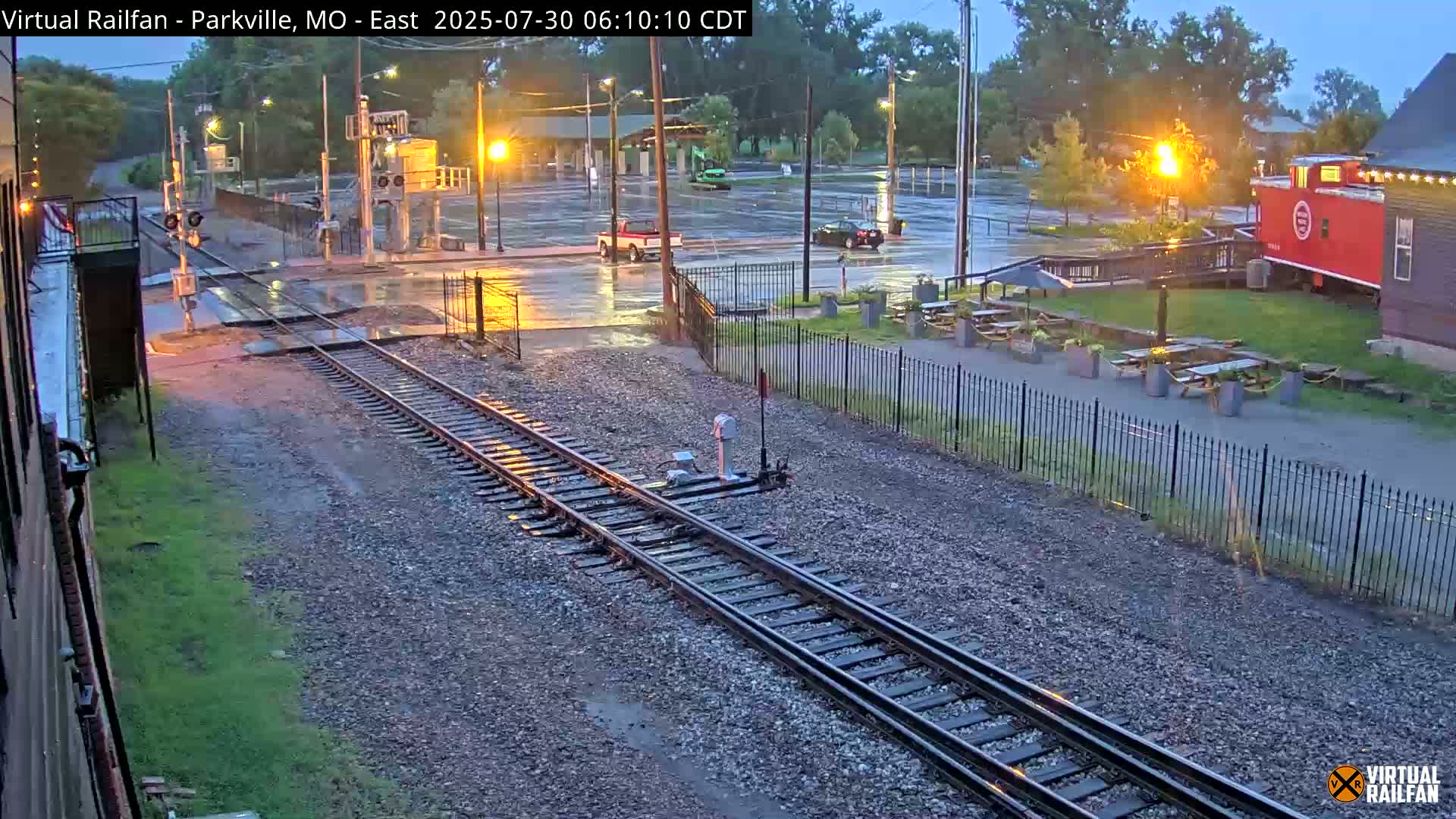 A nighttime view of a railway crossing near a building with outdoor seating, under partly cloudy skies.