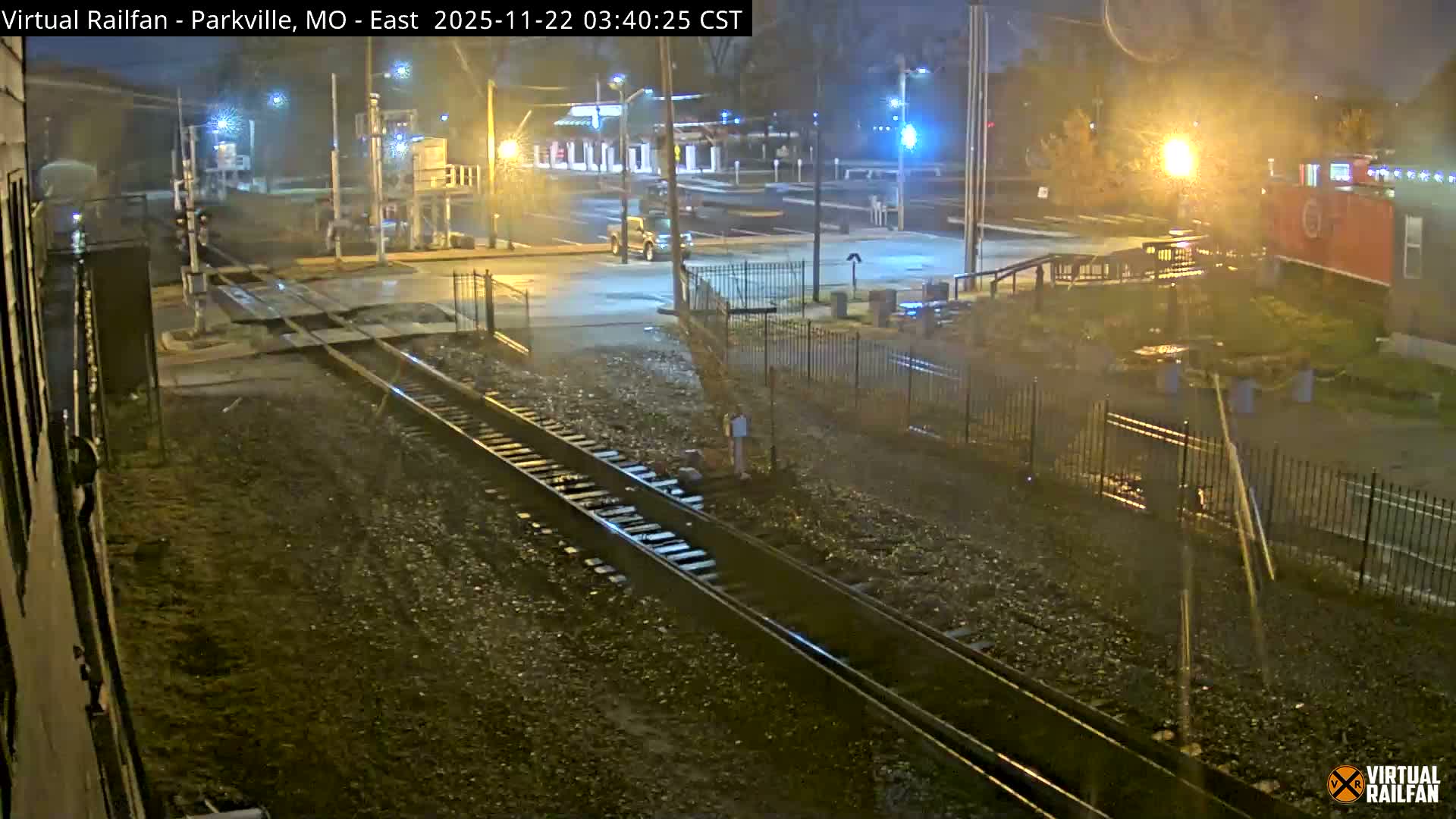 A wet railway crossing and adjacent road are illuminated by numerous streetlights at night, with train tracks prominently in the foreground and a vehicle visible on the road under misty or rainy conditions.