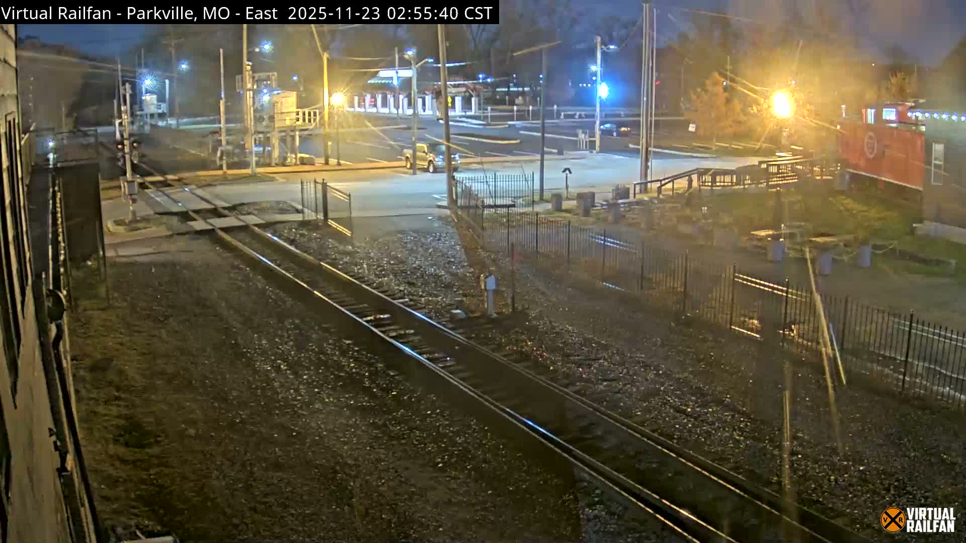 The image displays a nighttime scene of train tracks converging at a lighted road crossing, with buildings and a decorative red train car visible under artificial illumination in a clear, dark environment.